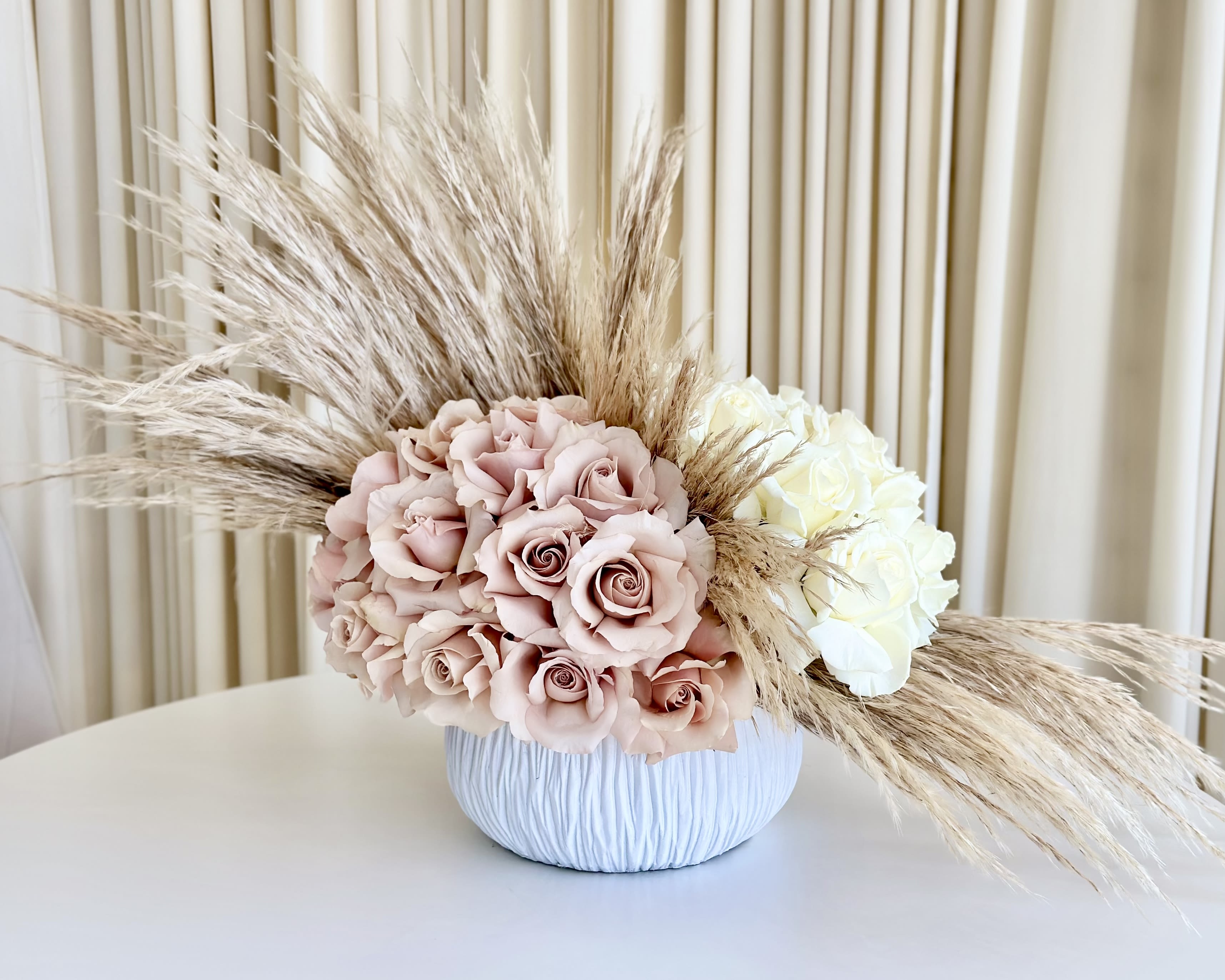 Whimsical Pampas Blush Whimsical Pampas Blush - Delight in the enchanting allure of "Whimsical Pampas Blush," a neutral flower arrangement that marries the elegance of pink and white roses with the ethereal beauty of pampas grass. Lovingly separated by the feathery texture of pampas, this captivating design is presented in an adorable and cute vase. Soft blush-pink roses exude grace and admiration, while pristine white roses symbolize purity and innocence. Together, they create a harmonious blend of colors, representing the delicate balance of emotions. The whimsical addition of pampas grass adds a touch of dreamy charm, evoking a sense of wonder and wanderlust. This natural accent brings a subtle, bohemian touch, making "Whimsical Pampas Blush" a unique and captivating arrangement that stands out in its simplicity.