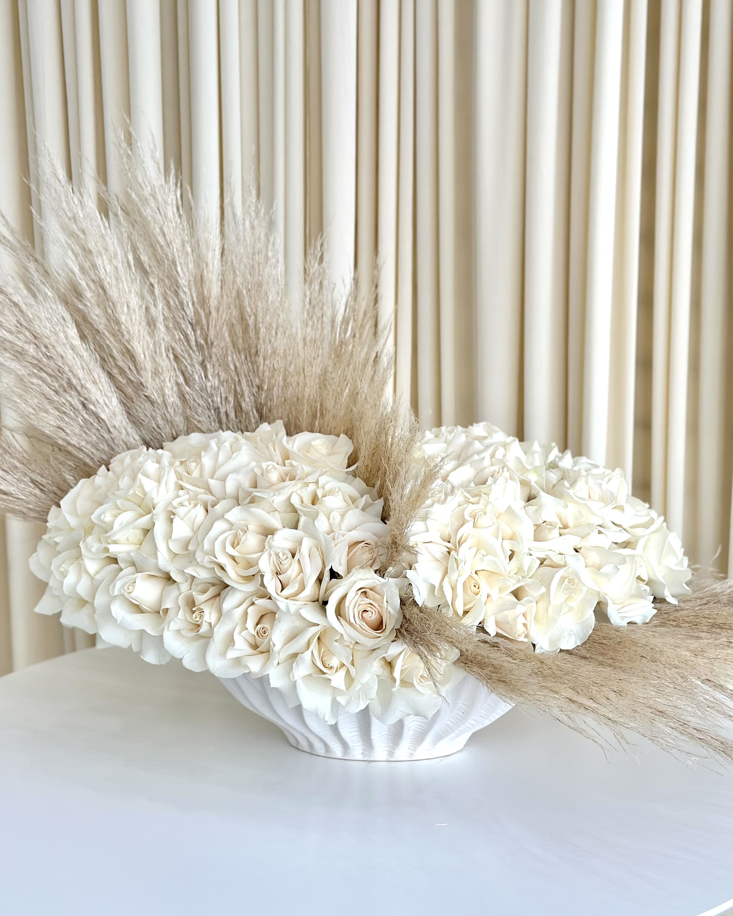 Whispering Serenity - Immerse yourself in the tranquility of &quot;Whispering Serenity,&quot; an exquisite all-white rose arrangement thoughtfully arranged in a beautiful white vase. With an elegant design accentuated by a line of delicate pampas grass, this masterpiece captures the essence of pure elegance and understated beauty.  Each white rose, representing purity and grace, finds its perfect place within the vase, forming a symphony of serenity and sophistication. The gentle whisper of the pampas grass adds a touch of ethereal charm, evoking a sense of calmness and poetic wonder.