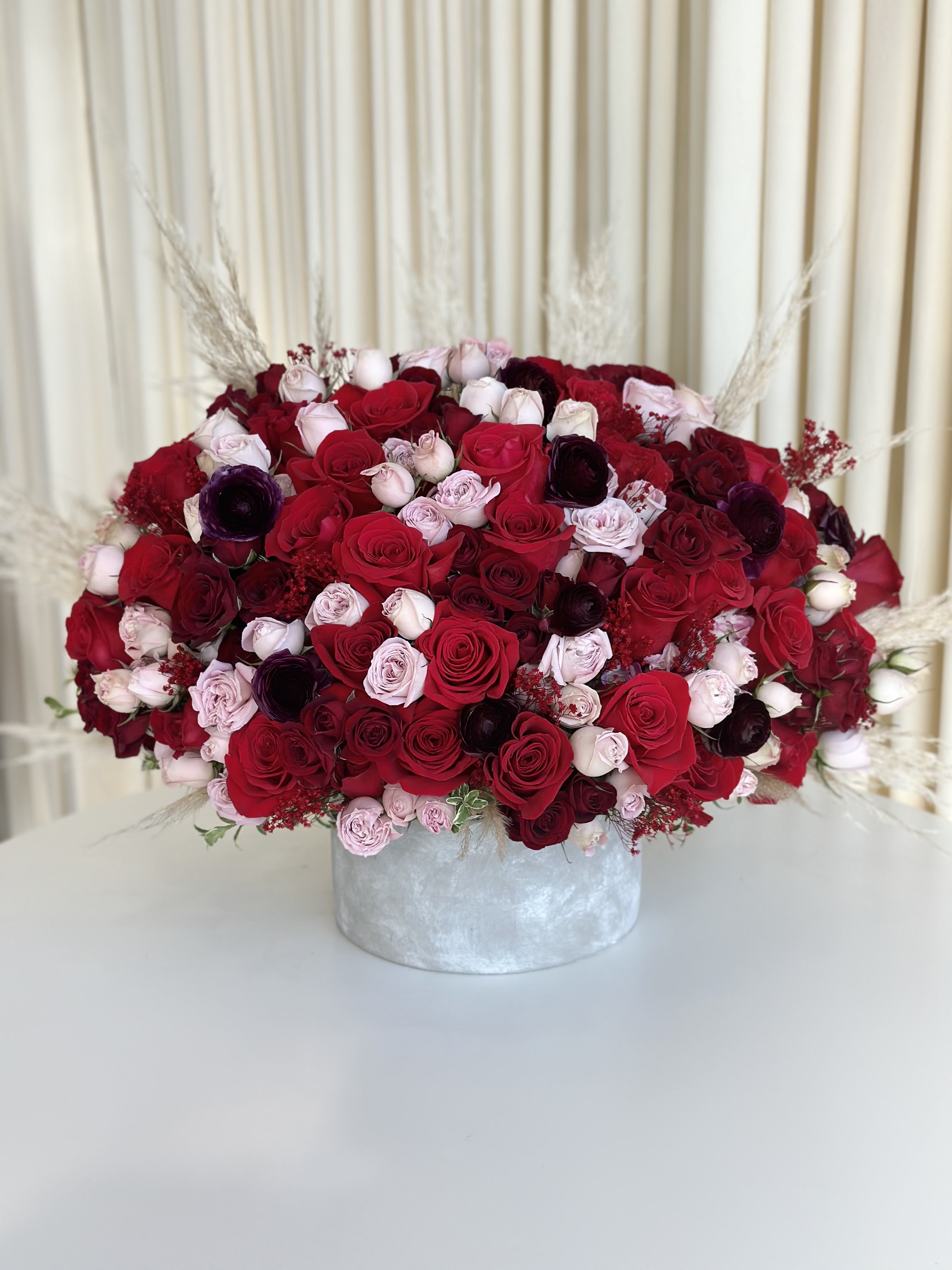 Enchanted Elegance - Elevate your emotions with our exquisite &quot;Enchanted Elegance.&quot; This stunning floral arrangement is a symphony of soft pink and bright red roses, deep red roses, deep purple ranunculus, and delicate dried florals thoughtfully intertwined to create a masterpiece that will captivate the heart.  The soft pink and bright red roses symbolize love, passion, and admiration, while the deep red roses add a touch of mystery and romance. The deep purple ranunculus brings a sense of enchantment and charm to the bouquet, making it truly unique.  Each petal is carefully selected and arranged to ensure a harmonious blend of colors and textures, creating a captivating visual and sensory experience. The accent dried florals add an artistic flair, making this bouquet a work of art that will be cherished and remembered.  Whether it's a declaration of love, a gesture of appreciation, or a heartfelt apology, the &quot;Enchanted Elegance&quot; speaks the language of the heart. Surprise your loved one with this enchanting arrangement and let the timeless beauty of roses convey your deepest emotions. 