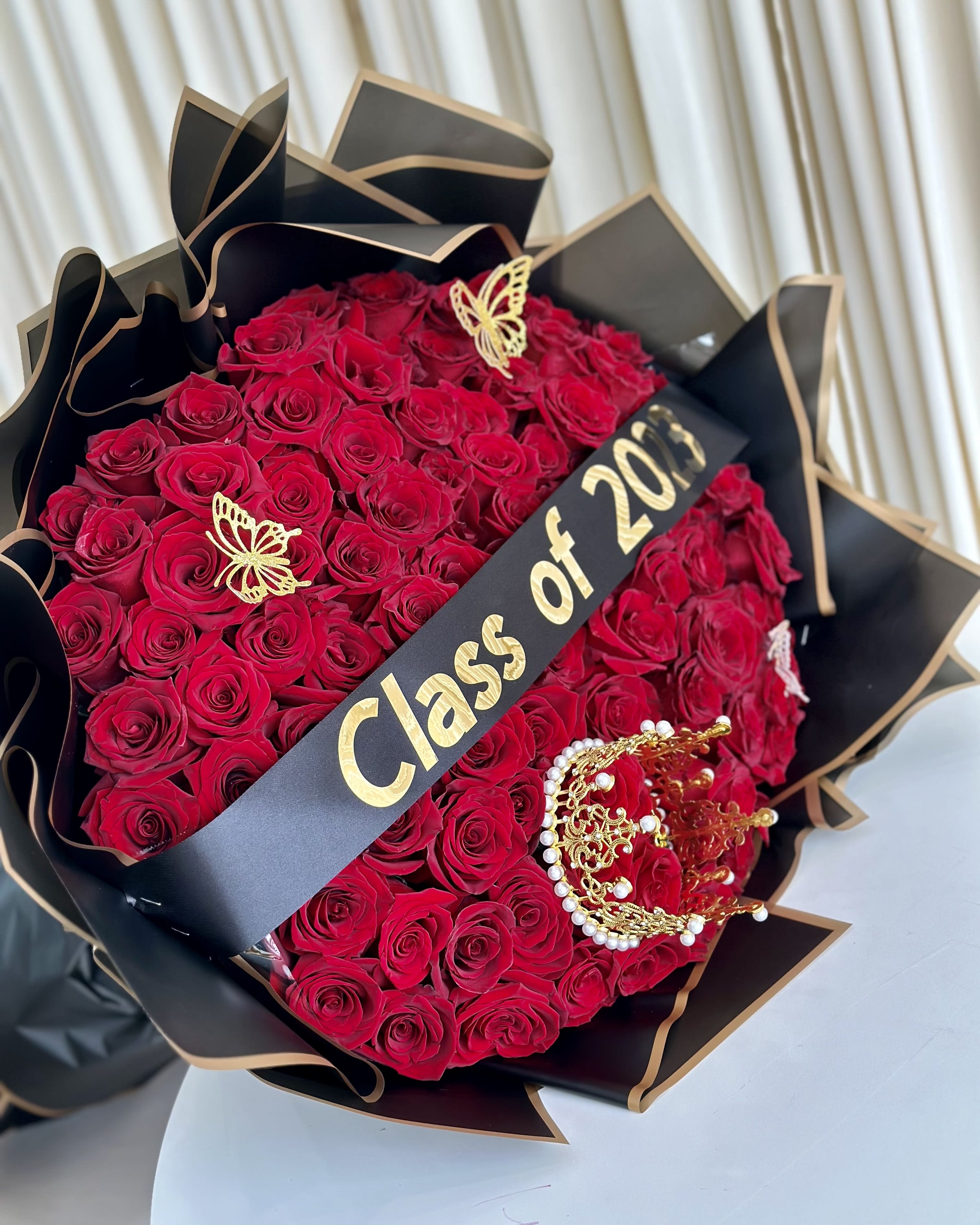 Eternal Achievement - Celebrate the journey of achievement with our &quot;Eternal Achievement&quot; Graduation Bouquet. This stunning arrangement features a bundle of vibrant red roses, each carefully selected to symbolize passion and success. Wrapped elegantly in black paper, adorned with gold accent butterflies and a crown, this bouquet exudes sophistication and triumph. Completing the look is a sash proudly displaying &quot;Class of 2023,&quot; commemorating this momentous milestone. Whether you're the graduate or want to congratulate someone special, &quot;Eternal Achievement&quot; is a heartfelt way to honor their accomplishments and create lasting memories. 