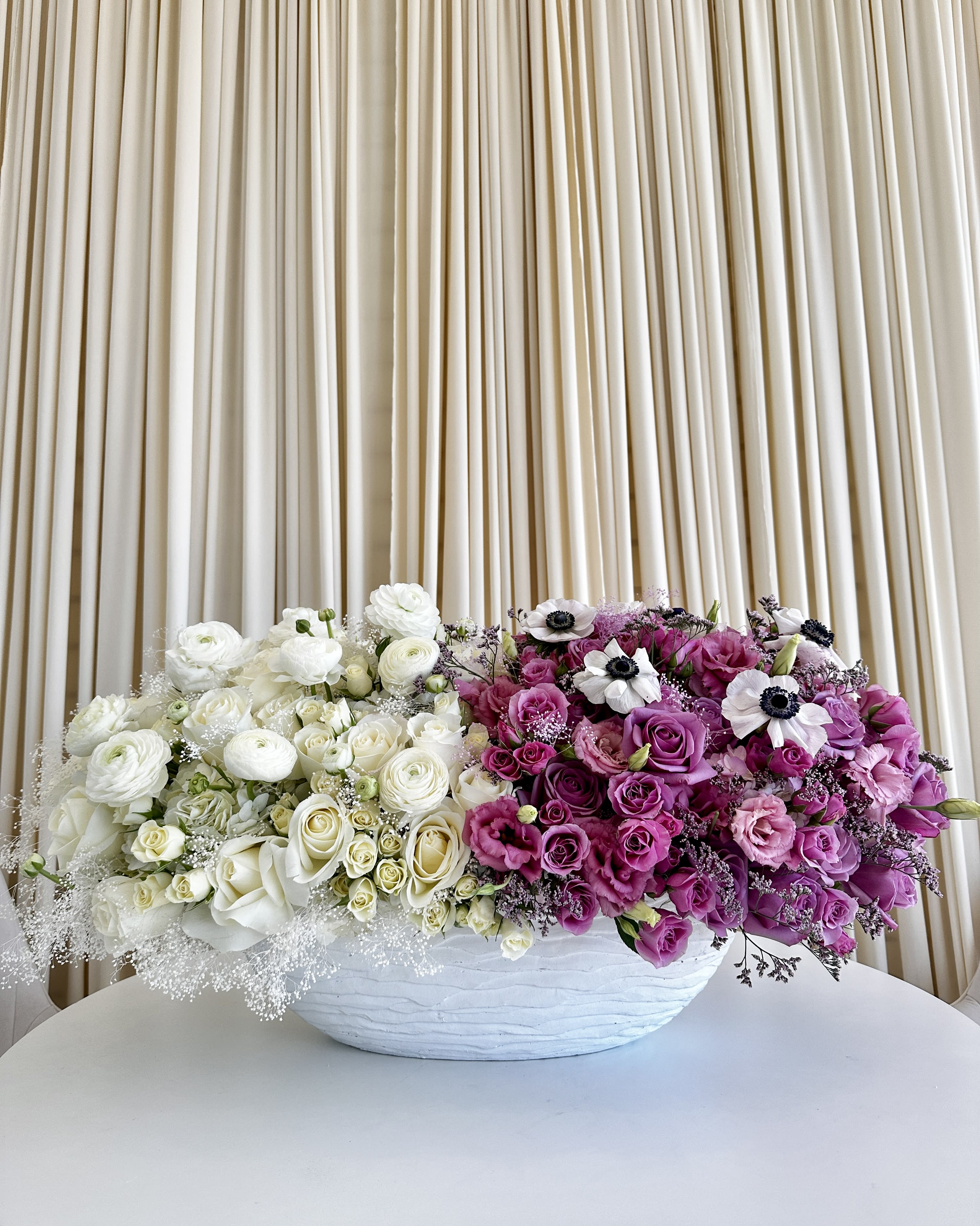 Harmony in Contrasts - Experience the exquisite &quot;Harmony in Contrasts&quot; vase arrangement, where the timeless elegance of all-white florals meets the enchanting allure of all-purple blooms. This stunning centerpiece showcases a delicate balance of beauty and sophistication, featuring pristine roses, elegant anemones, charming ranunculus, and graceful lisianthus flowers.  One side of the large white vase boasts a symphony of pure white blooms, radiating a sense of purity and grace. On the other side, a mesmerizing display of rich purple hues evokes a sense of mystery and fascination. Together, they create a captivating juxtaposition that celebrates the beauty of both ends of the floral spectrum.  Whether it's adorning your home, gracing a special occasion, or expressing your heartfelt sentiments to a loved one, the &quot;Harmony in Contrasts&quot; arrangement is an elegant embodiment of floral artistry. Embrace the harmony of two worlds, elegantly united in this distinctive creation. 