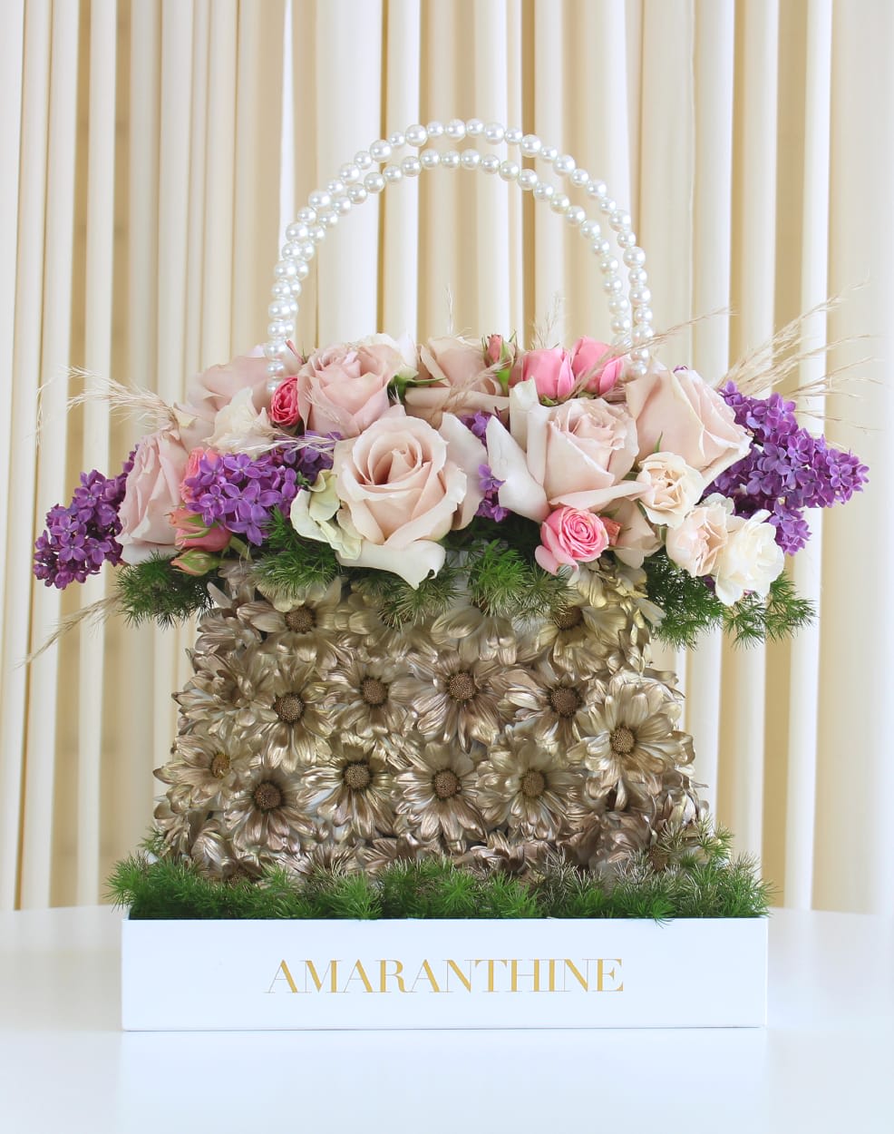 Golden Floral Purse - Elevate your floral gifting to new heights with our enchanting &quot;Golden Floral Purse.&quot; This unique flower arrangement is a delightful masterpiece crafted to resemble a stylish purse. The base boasts glistening gold-sprayed daisies, adding a touch of glamour to the design. Adorned with beautiful pink roses and elegant purple lilacs at the top, this purse-inspired arrangement is a perfect blend of sophistication and natural beauty. Surprise your loved ones with this charming gift that's sure to leave a lasting impression. Express your affection with a purse that will never go out of style - a purse filled with exquisite blossoms and heartfelt sentiments. 