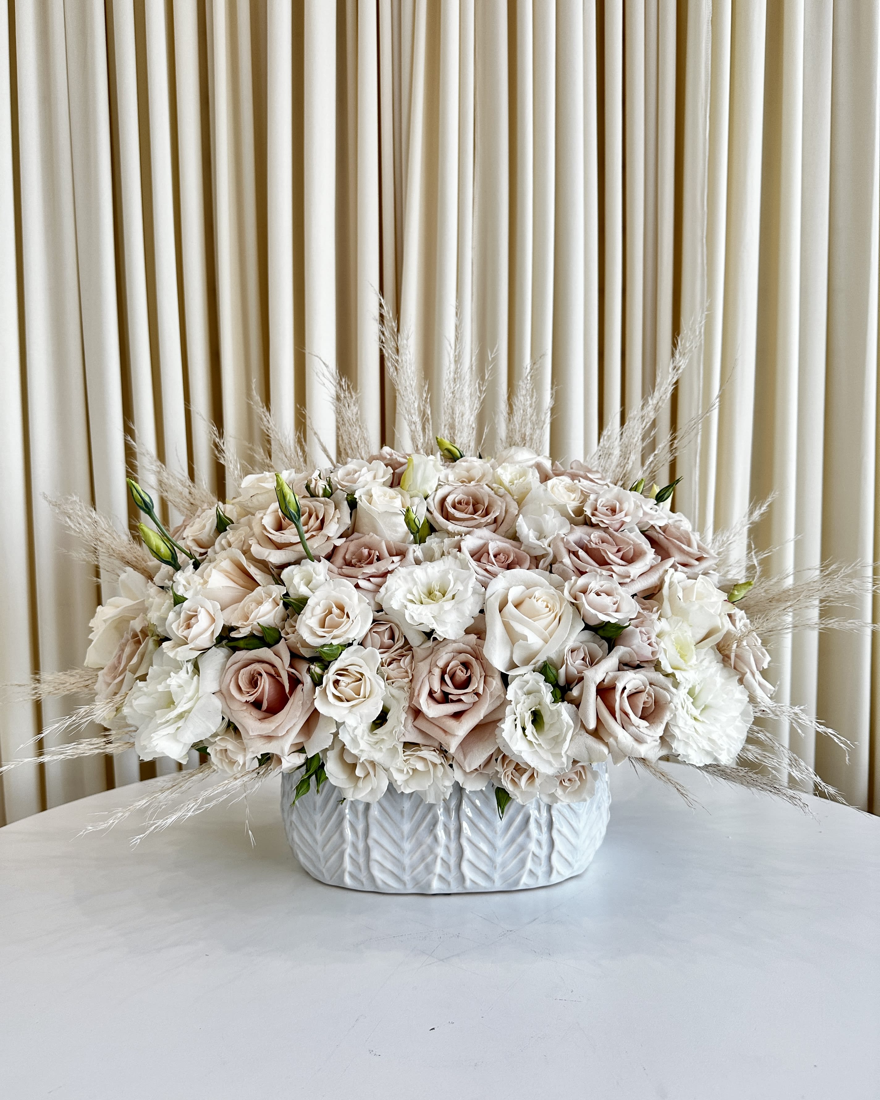Serenity In Neutrals - !!! This vase is discontinued. We will substitute it for a similar vase !!! Immerse yourself in the tranquility of &quot;Serenity in Neutrals&quot; – a captivating floral arrangement designed to bring calm and elegance to any space. Nestled in a beautiful white vase, this exquisite creation features a harmonious palette of lisianthus and roses in soothing neutral hues, complemented by a gentle touch of pampas grass.  Delicate lisianthus blooms dance gracefully alongside the timeless beauty of roses, creating a harmonious symphony of textures and colors. The neutral tones add an air of sophistication and refinement, making this arrangement a versatile and elegant addition to any setting.  The subtle inclusion of pampas grass adds a touch of natural charm, invoking a sense of peacefulness and harmony with nature. &quot;Serenity in Neutrals&quot; is more than just a floral arrangement; it's an invitation to embrace the serene beauty that blooms bring into our lives.  Whether you're looking to create a serene ambiance at home, gift a loved one with a heartfelt sentiment, or adorn a special event with understated elegance, &quot;Serenity in Neutrals&quot; is the perfect choice. Elevate your surroundings with this enchanting masterpiece and let the soothing tones of nature's finest blooms whisk you away to a world of calm and serenity.