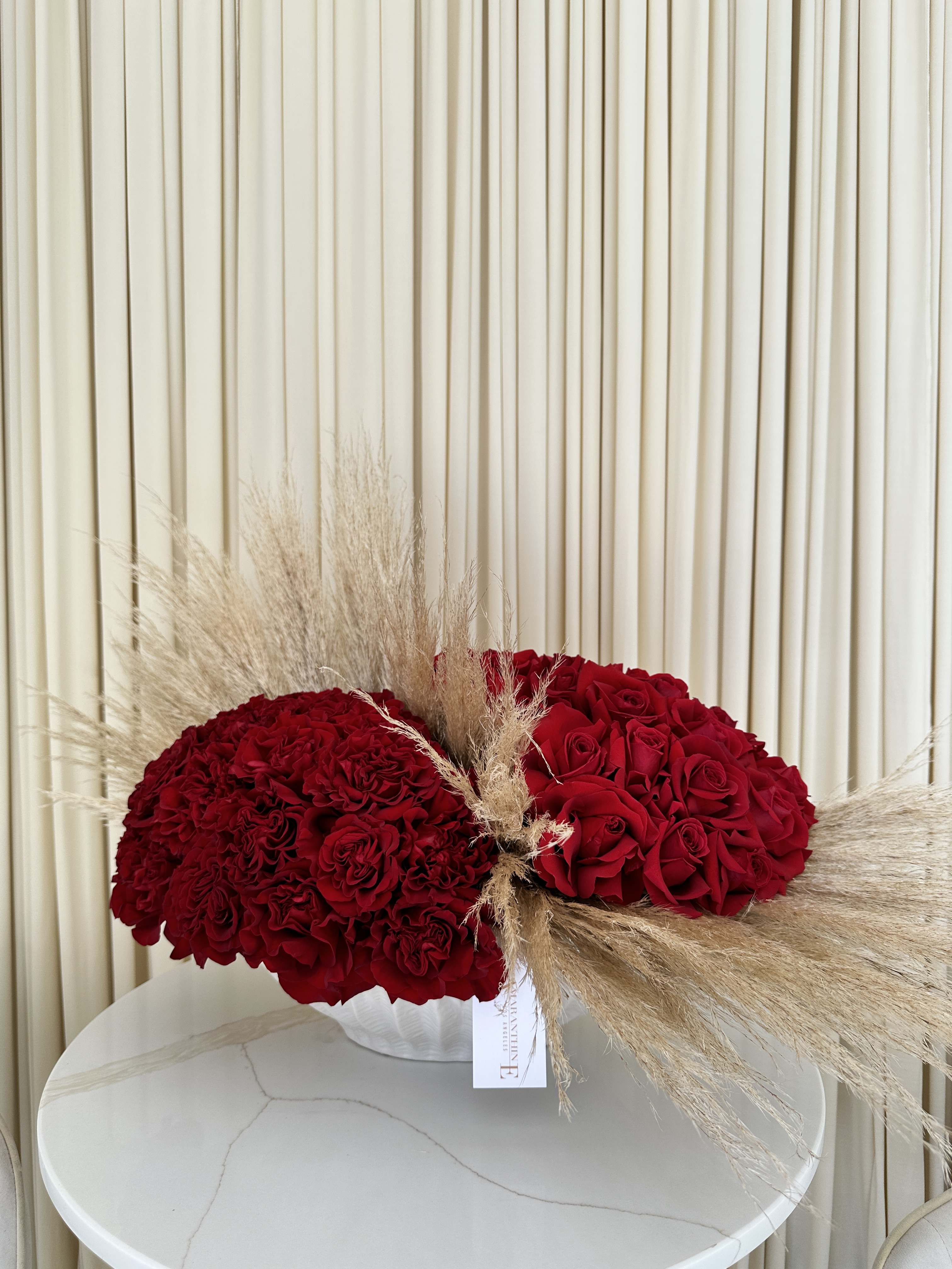 Whispers of Red - Enhance the ambiance of any space with our exquisite red rose and pampas grass arrangement. The vibrant red roses symbolize love and passion, while the delicate pampas grass adds a touch of elegance and sophistication. Carefully arranged in a stylish vase, this stunning floral arrangement is perfect for adding a pop of color and natural beauty to your home or office. Whether you're looking to impress someone special or simply want to treat yourself, this red rose and pampas grass arrangement is sure to make a lasting impression.