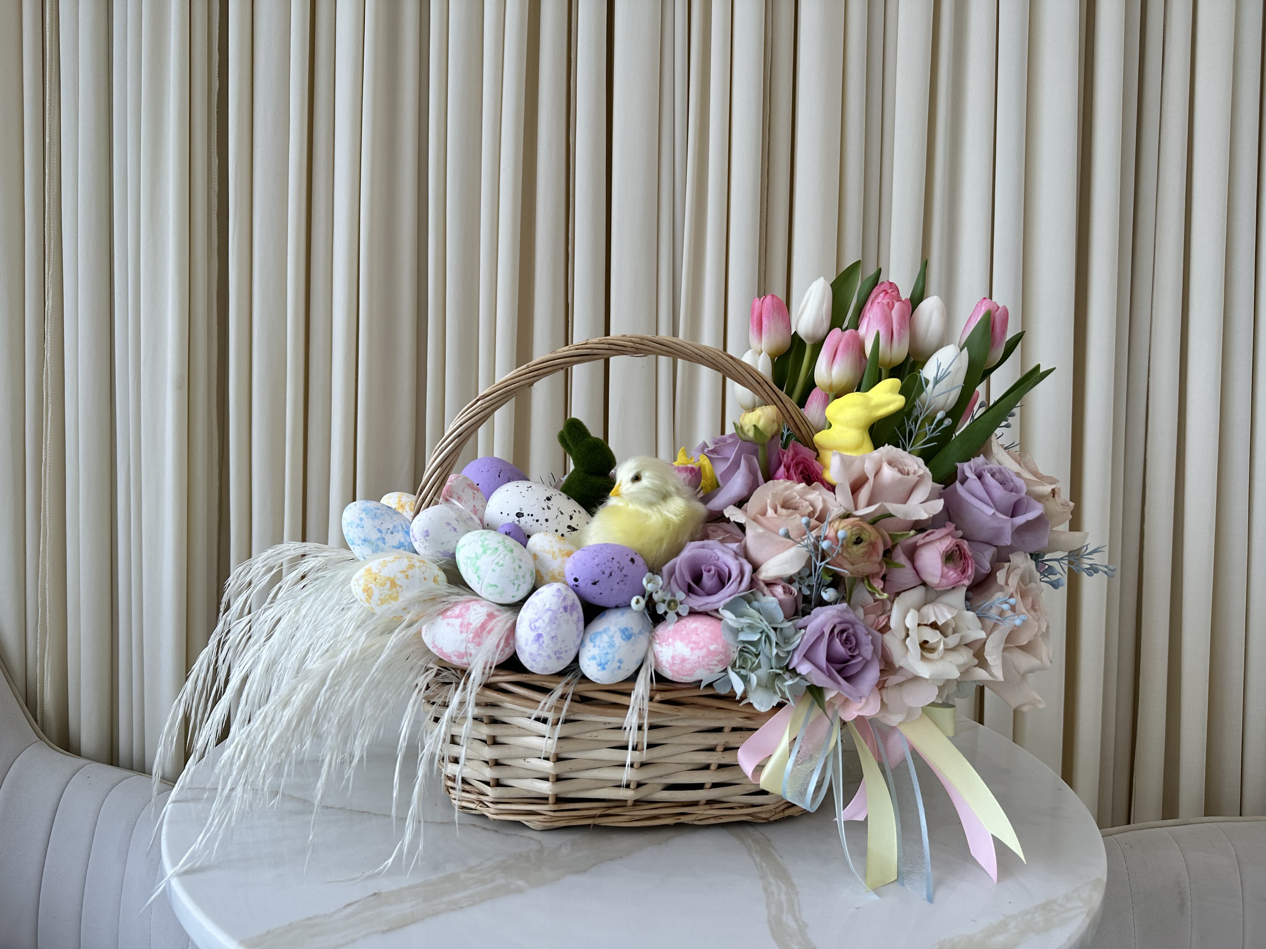 Easter Chick - Celebrate the joy of Easter with this delightful basket filled with springtime blooms and a touch of Easter cheer. This charming arrangement features pink and purple roses, and a fluffy chick figurine, all nestled together in a beautiful woven basket. Perfect for brightening someone's day or as a festive centerpiece for your Easter celebration.