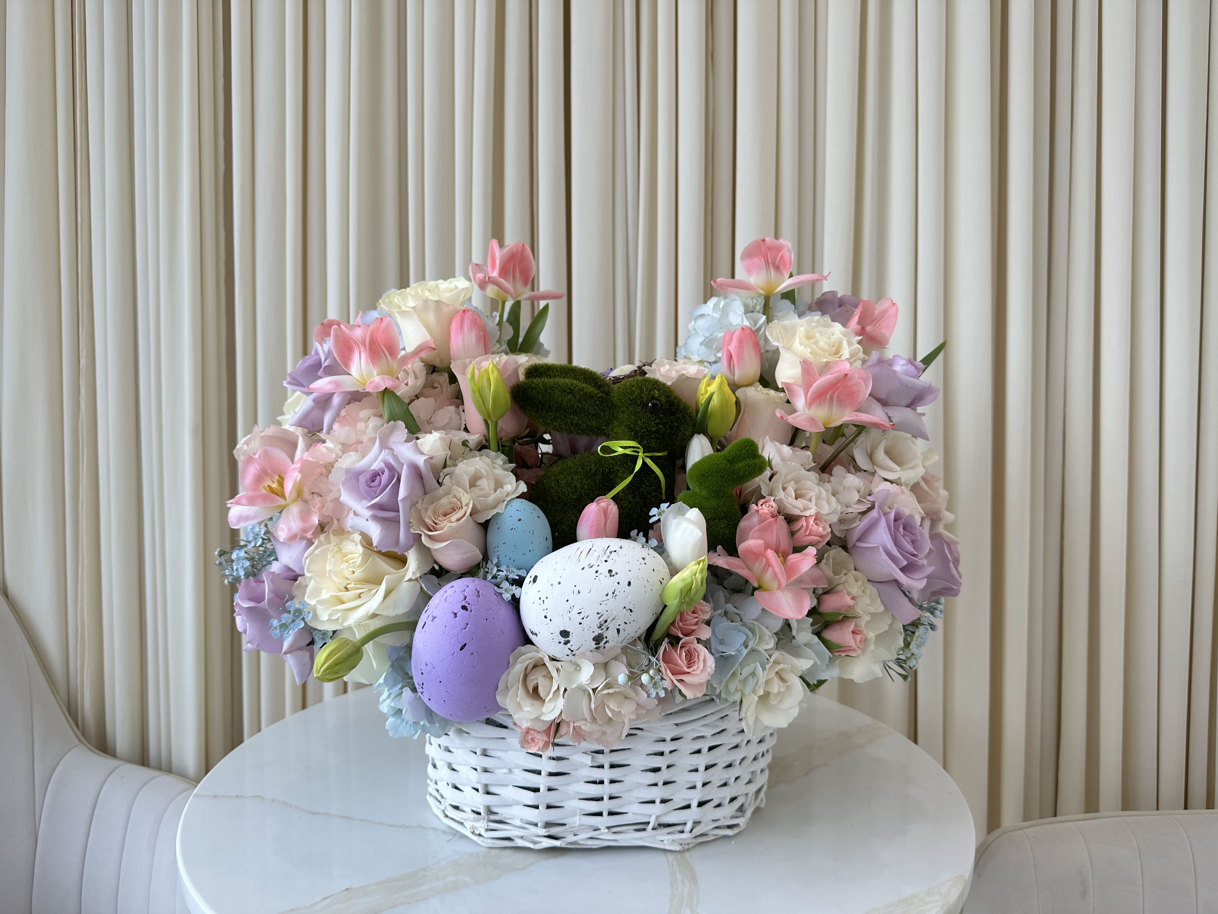 Easter Sunshine - Welcome the Easter season with this delightful basket overflowing with pastel-colored blooms and a touch of springtime cheer. Featuring pink and yellow tulips, roses in soft pastel hues, this basket is accented with a sweet bunny figurine, creating a charming way to celebrate new beginnings.
