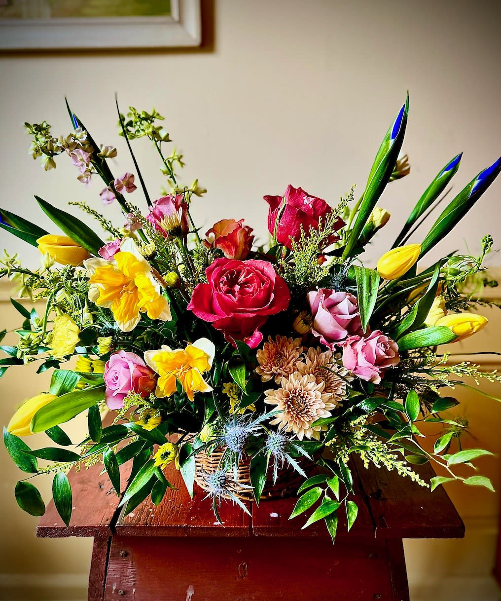 Family Woven - The arrangement is set in a rustic woven basket, brimming with a lush assortment of roses and seasonal flowers. The roses, in shades of soft pink, creamy white, and deep red, form the focal point, their velvety petals creating a sense of timeless elegance. Surrounding the roses are seasonal blooms, such as bright sunflowers, delicate dahlias, and vibrant zinnias, adding bursts of color and texture. Sprigs of fresh greenery, like eucalyptus and ferns, weave through the arrangement, giving it a natural, garden-like feel. A few delicate tendrils spill over the edges of the basket, adding a touch of whimsy and charm. The overall effect is one of abundant beauty, perfect for brightening any space.