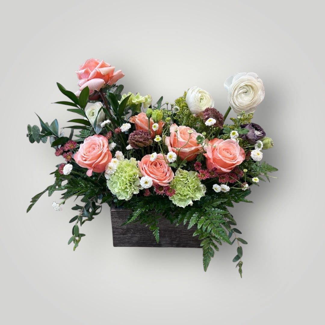 Noah Noah - his floral arrangement is a rectangular, garden-inspired display featuring a mix of soft peach and white tones with lush greenery. All of these are displayed in a rustic, dark wooden container that complements the organic and vibrant feel of the flowers. This arrangement is perfect for a cheerful, elegant setting.