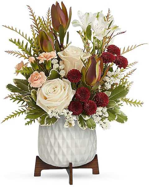 Timeless Treasure Bouquet - Showcasing radiant red and cream blooms presented in a sleek geometric ceramic planter atop a retro-inspired wooden base. It's an inspiring blend of elegance and nostalgia. Experience refined beauty with a bouquet of cream roses, white alstroemeria, red leucadendron, peach carnations, and burgundy chrysanthemums. Accented by white sinuata statice, grevillea, and variegated pittosporum, this elegant arrangement is presented in the Mid Mod Planter, epitomizing sophistication and style.  