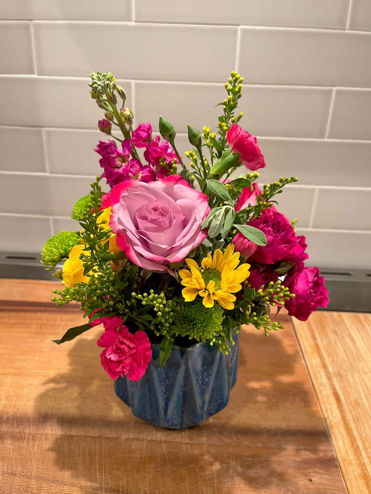 Begin Again - A vibrant mix of cool lavender and Pink Floyd roses. crowned with bright pink stock flower, spray roses and pops of yellow pom daisies. A wonderful way to bring a smile to someone's face!  These work great as a gift, table centerpiece, or event decor. If ordering for an event, 3-4 arrangements work best for a table of 12 guests. 