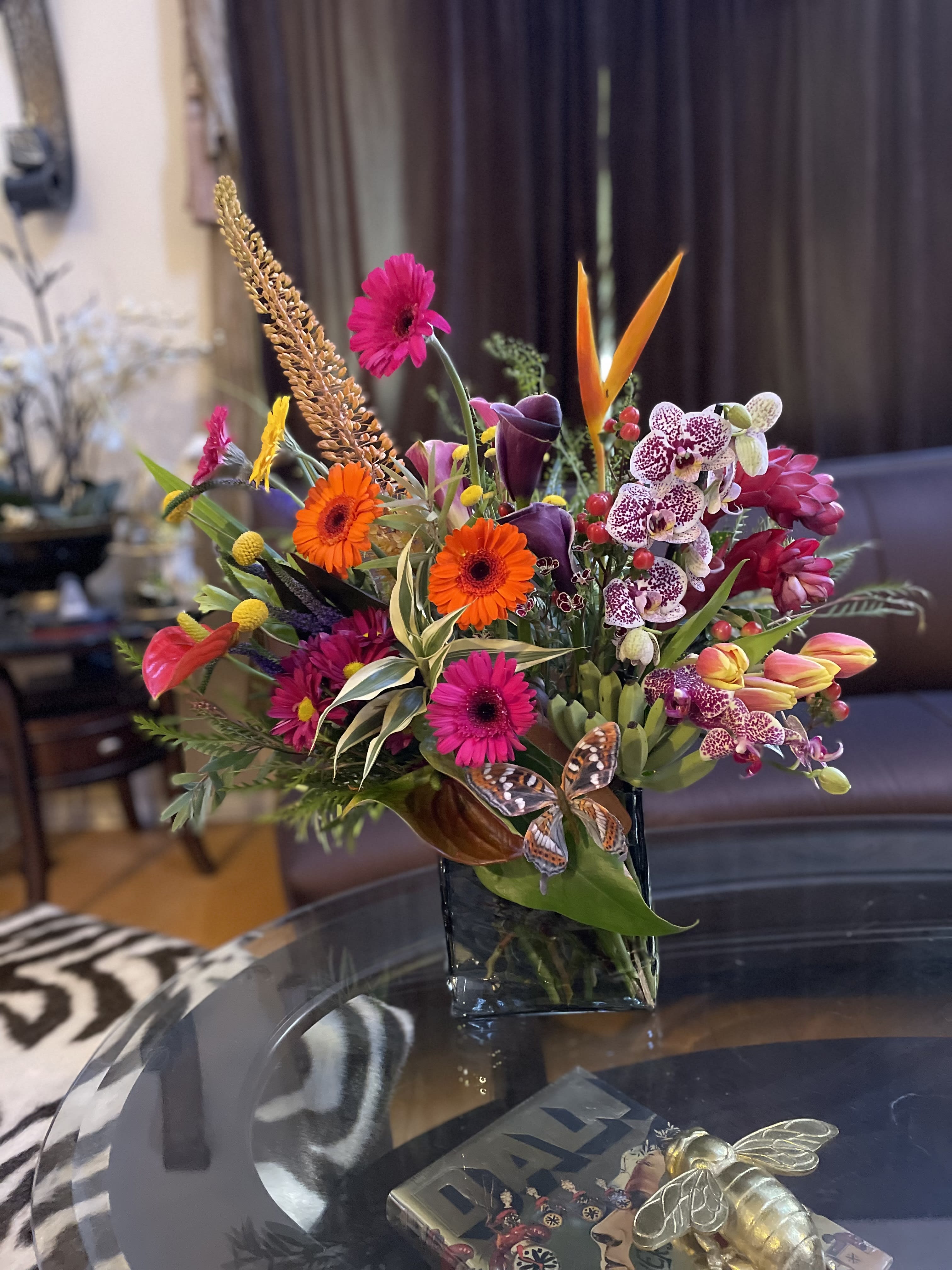 Tropical fusion  - A vibrant and exotic fusion of tropical elements creates an awe-inspiring arrangement that transports you to a paradisiacal oasis. Green leaves and baby bananas , fiery orange heliconias, velvety red anthuriums, and bold orange and magenta gerbera daisies intertwine with cascading green vines, while vibrant orchids, tulips, and calla lilies add a touch of elegance. The arrangement bursts with intense colors, reminiscent of a tropical sunset, and the intoxicating scent of tropical ginger blooms fills the air. This tropical fusion arrangement effortlessly captures the essence of a tropical paradise, bringing a sense of warmth, relaxation, and adventure to any space. Great gift for your Boss !