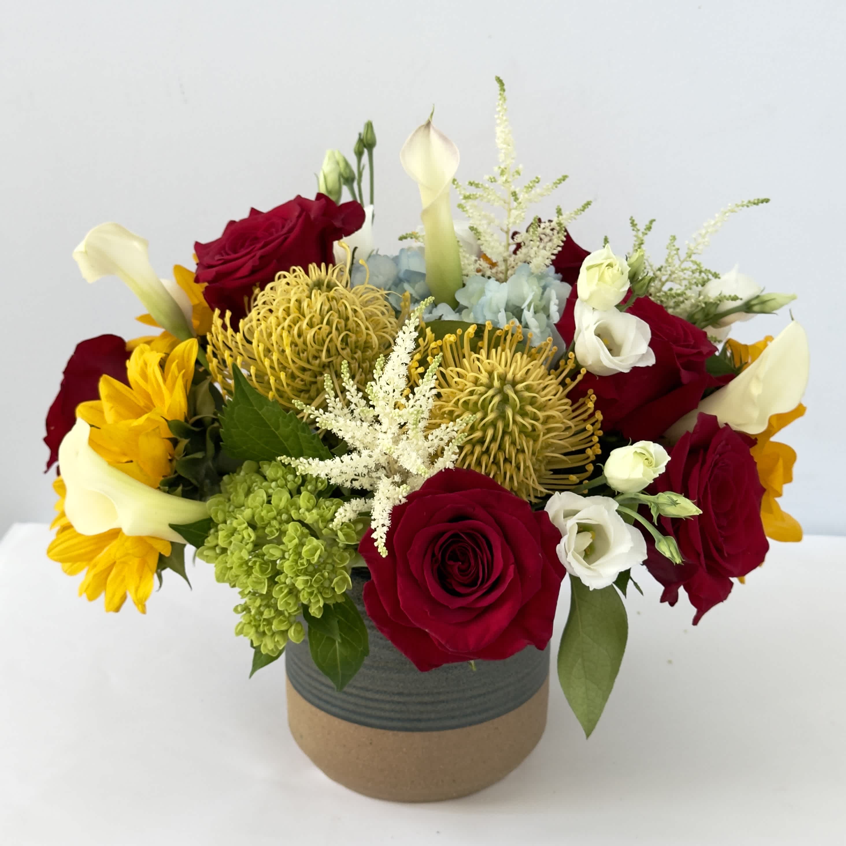 Carmine Carmine - This arrangement is presented in a 5" ceramic vase. It features sunflowers, red roses, yellow pincushions and blue hydrangea.