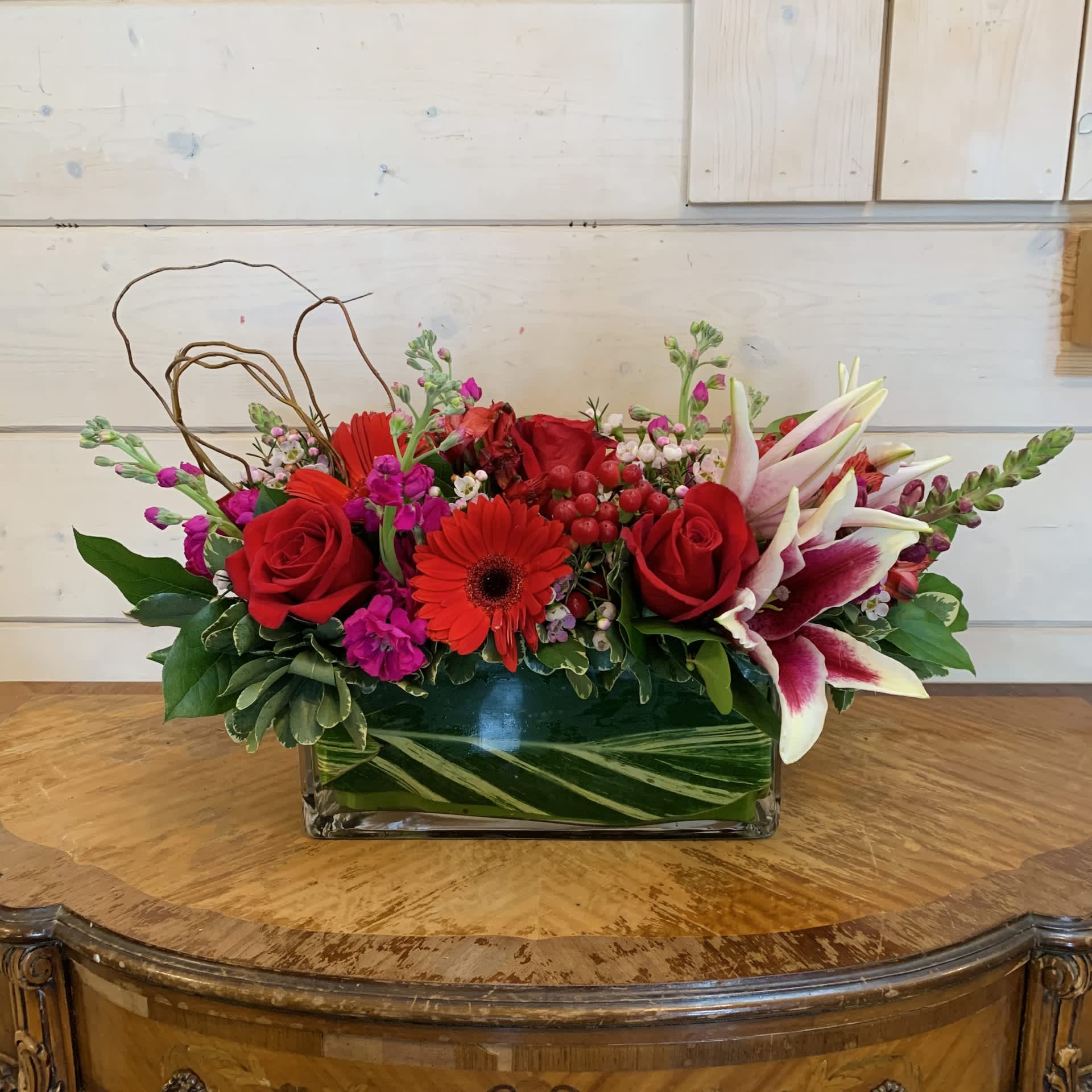 Pure Romance Centerpiece Pure Romance Centerpiece - Red roses, stargazer lily, red gerbera daisies, red hypericum, magenta stock, burgundy snaps and red alstroemeria arranged in a 12 inch leaf lined long and low glass vase with seasonal filler flower and looped curly willow. Approximately 17" long, 9" high and 8" wide.