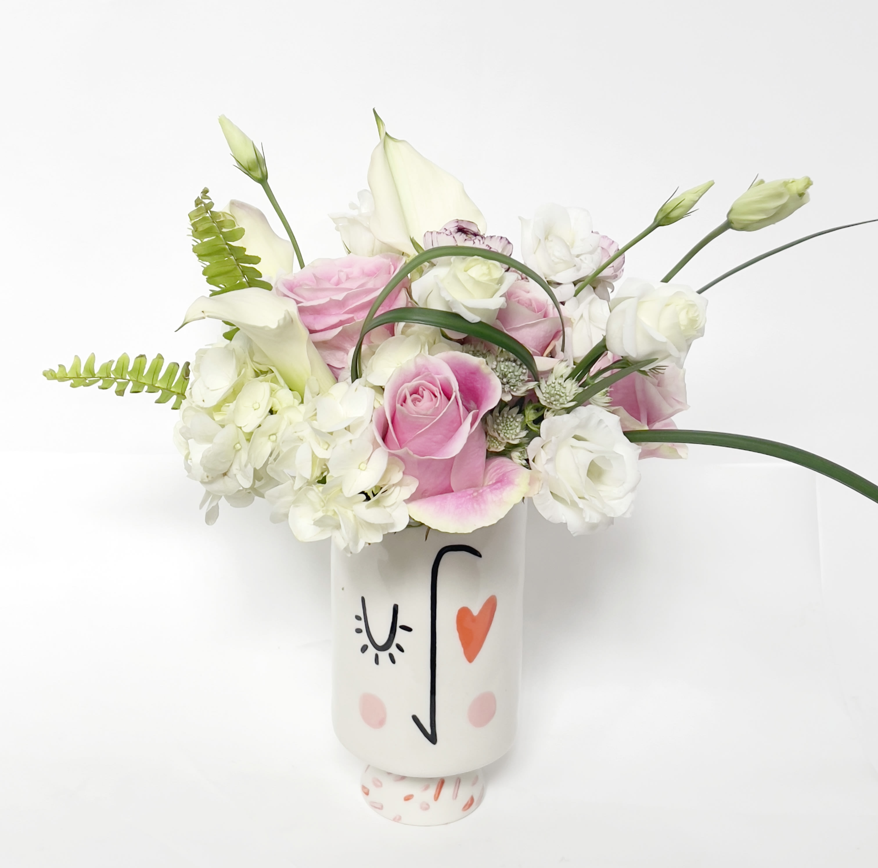 Heart Eye  - This is an elegant and fun floral design, inspired by its vessel – a modern white ceramic vase with a wink and a heart. Beautiful pink roses and bicolor ranunculus pop amidst the classic whites (hydrangeas, calla lilies, lisanthus) and subtle green accents.