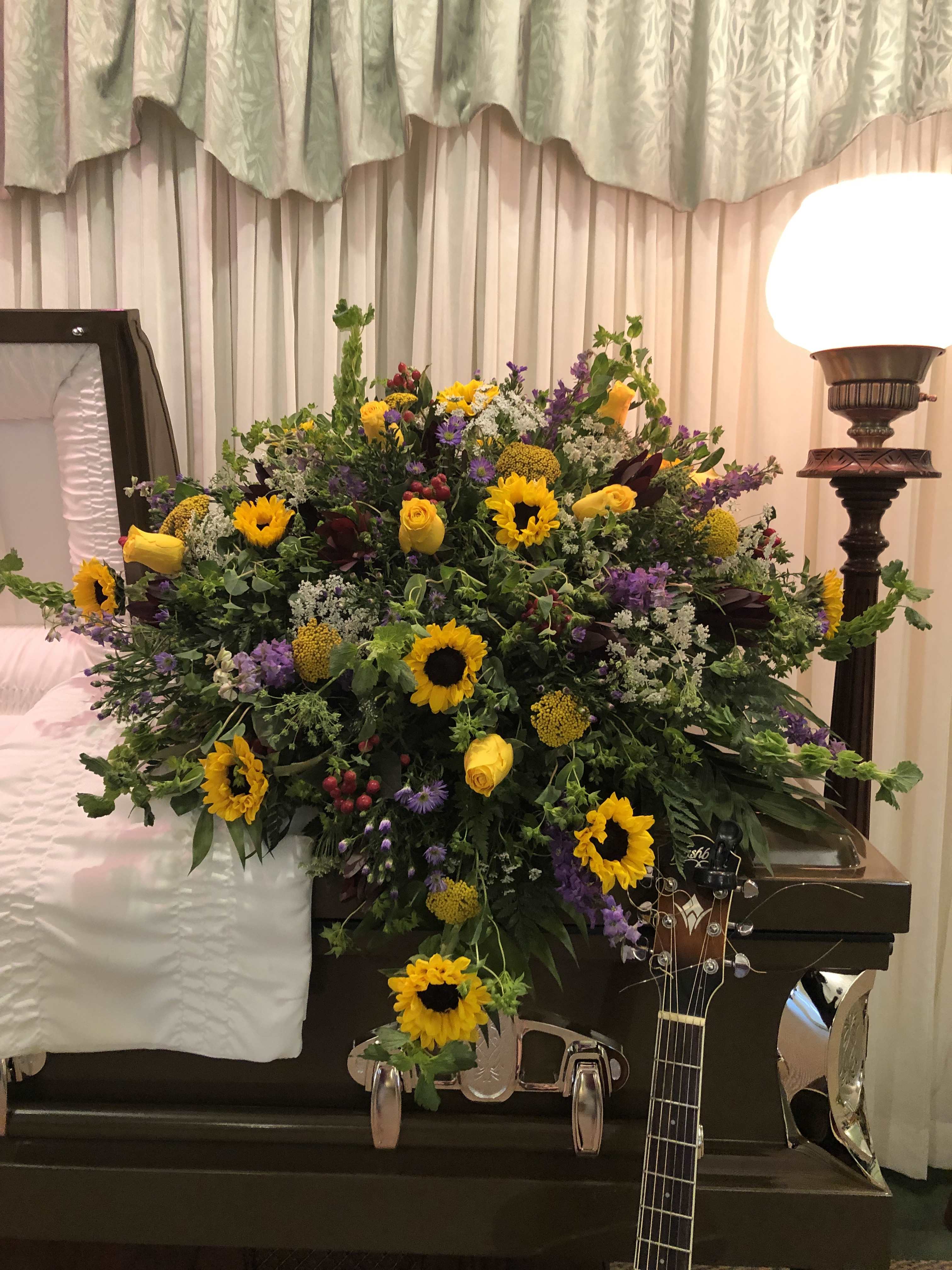 Sunflower Garden Casket Spray - Sunflowers, yellow roses, safari sunset, purple larkspur, queen Ann’s lace, yellow yarrow, red berries, bell of Ireland, and purple monte casino with blupernum, emerald, and leather leaf designed in a traditional cascade style.
