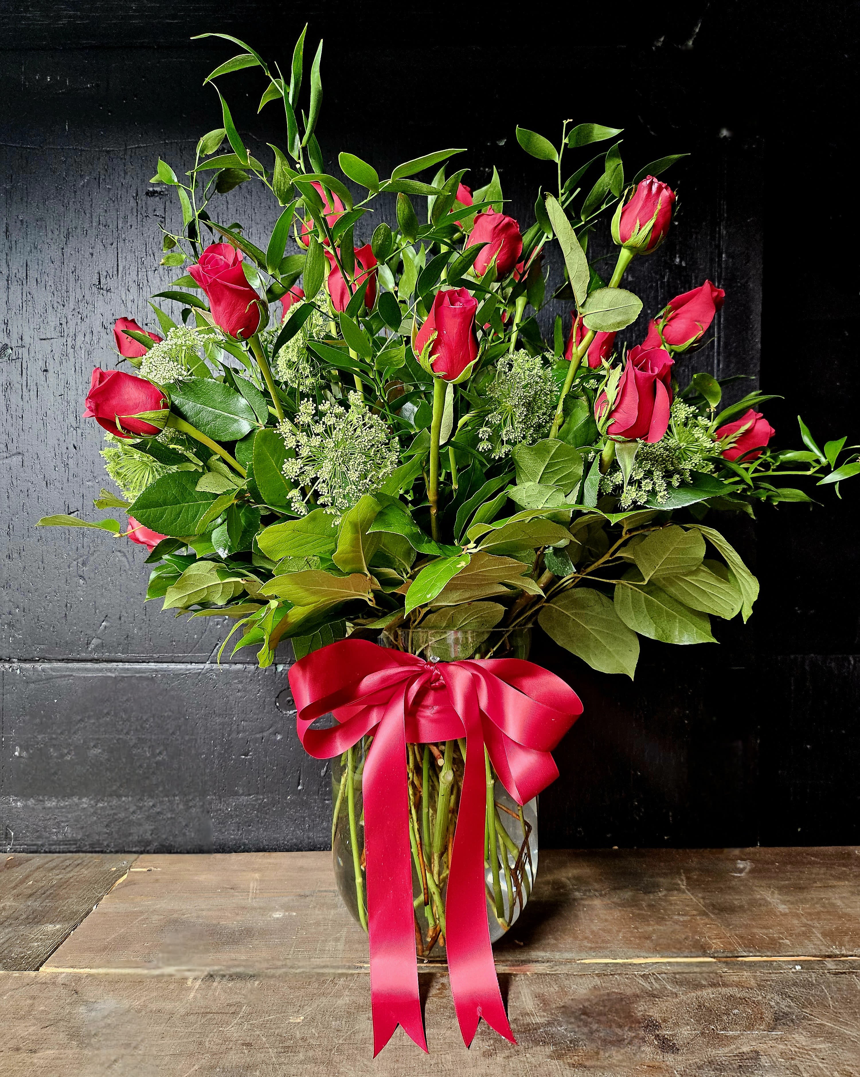 Elegant Red Rose Arrangement - Celebrate love, beauty, and elegance with this timeless arrangement of stunning red roses, complemented by lush greenery and delicate Queen Anne's Lace for added sophistication. The roses are artfully arranged in a clear glass vase and adorned with a luxurious red satin bow, making it the perfect gift for anniversaries, romantic occasions, or simply to brighten someone's day.  This arrangement is a testament to classic charm and heartfelt sentiments, ideal for expressing love and admiration.  Care Instructions:  1. Keep in a cool location, away from direct sunlight and heat sources.   2. Refill the vase with fresh water daily to maintain hydration.   3. Trim the stems every 2–3 days at an angle for optimal water absorption.