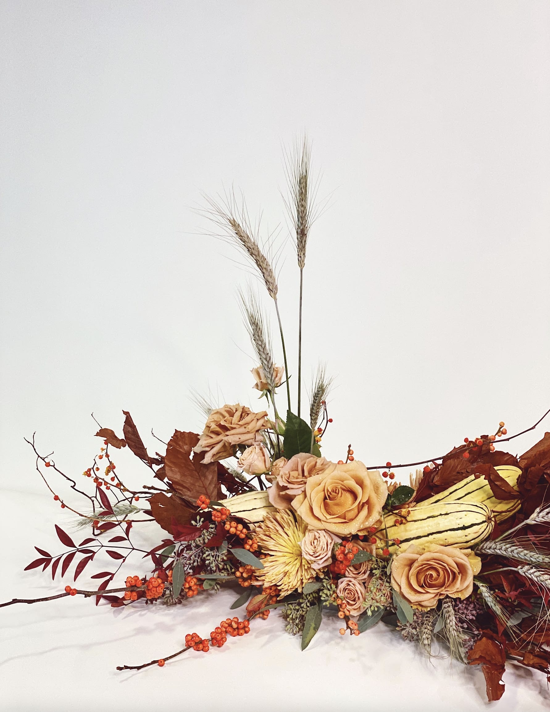 WHEAT-SCAPE - - Stunning harvest-themed arrangement - Features gold-toned roses, vibrant orange berries, rustic wheat stalks, and festive gourds - Ideal centerpiece for family gatherings or welcoming guests - Celebrates the season with charm