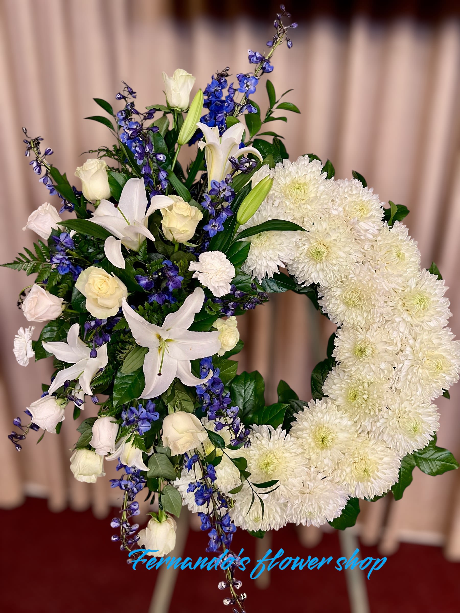 Blossoms of Confort - This elegant floral arrangement features a striking wreath design crafted with pure white chrysanthemums, symbolizing peace and purity. Accentuating the design are graceful white lilies, delicate white roses, and vibrant blue delphiniums, adding depth and a serene pop of color. Lush greenery provides texture and balance, enhancing the overall visual appeal. Perfect for honoring a loved one’s memory, this arrangement exudes grace, dignity, and a heartfelt tribute to life. Created with care, it reflects a message of remembrance and peace.