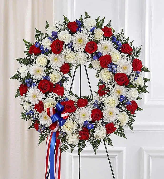 Freedon  - This patriotic wreath is a heartfelt tribute to honor and remember a loved one who served with courage and dedication. Featuring a stunning combination of red roses and carnations, white chrysanthemums, and blue delphiniums, this arrangement is a symbol of pride and gratitude. Accented with lush greenery and adorned with a red, white, and blue ribbon, it beautifully expresses respect and admiration. Perfect for memorial services or commemorative events, this wreath stands as a proud salute to their legacy.