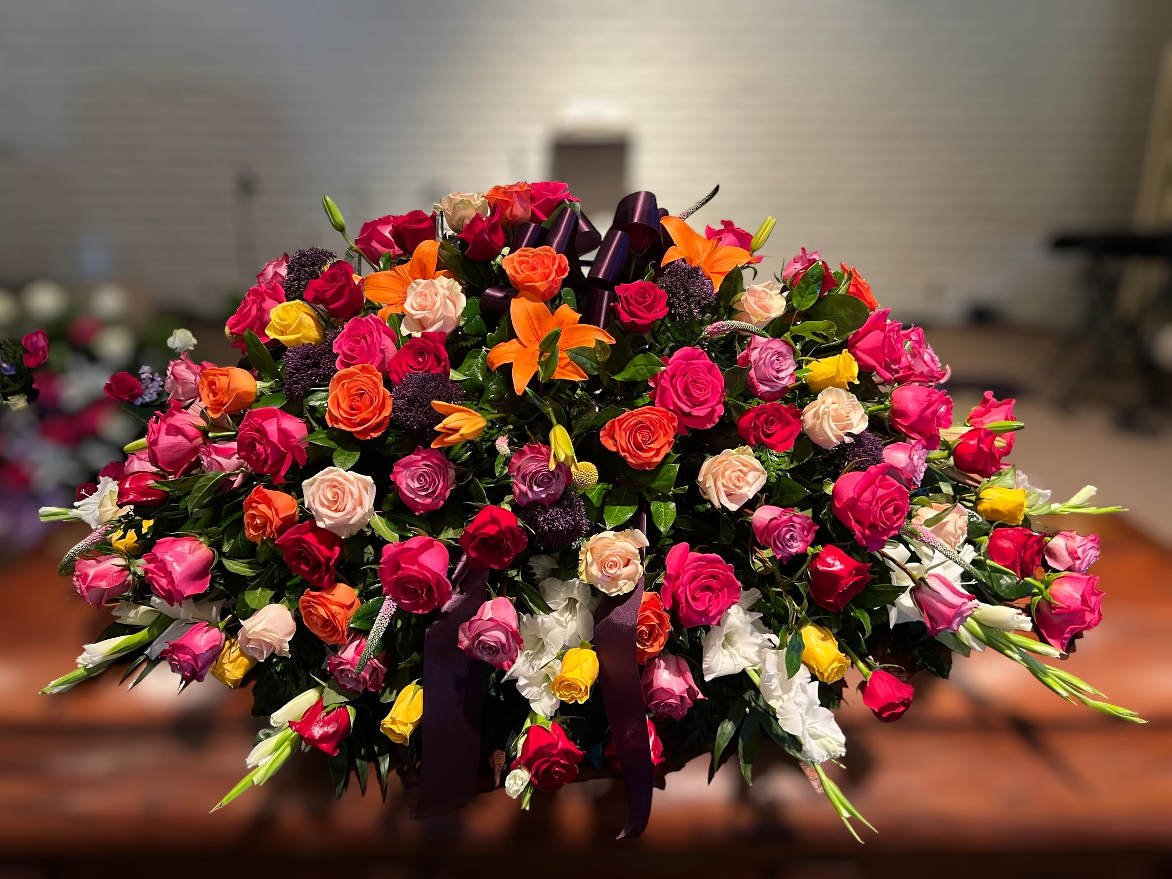 Petals of Peace By Fernando’s Flower Shop - This flower arrangement is a stunning and vibrant display designed to make an impactful statement. It is a large, full casket spray arrangement, commonly used for memorial or funeral services. Key features include: 	•	Roses in a variety of colors: Pink, red, orange, peach, yellow, and white roses dominate the design, creating a dynamic mix of warm and soft tones symbolizing love, remembrance, and admiration. 	•	Orange lilies: These bright and dramatic blooms add a focal point with their bold color and unique shape. 	•	Purple flowers (possibly scabiosa or similar): Providing depth and contrast against the brighter hues, enhancing the arrangement’s richness. 	•	White gladiolus: Tall and elegant, adding structure and height to the composition. 	•	Greenery: Lush leaves and filler greens create a fresh and balanced backdrop for the colorful flowers.  The arrangement is accented with a dark purple ribbon, adding a touch of elegance and solemnity. Its large size and colorful palette make it a perfect choice for honoring and celebrating a loved one with beauty and grace.