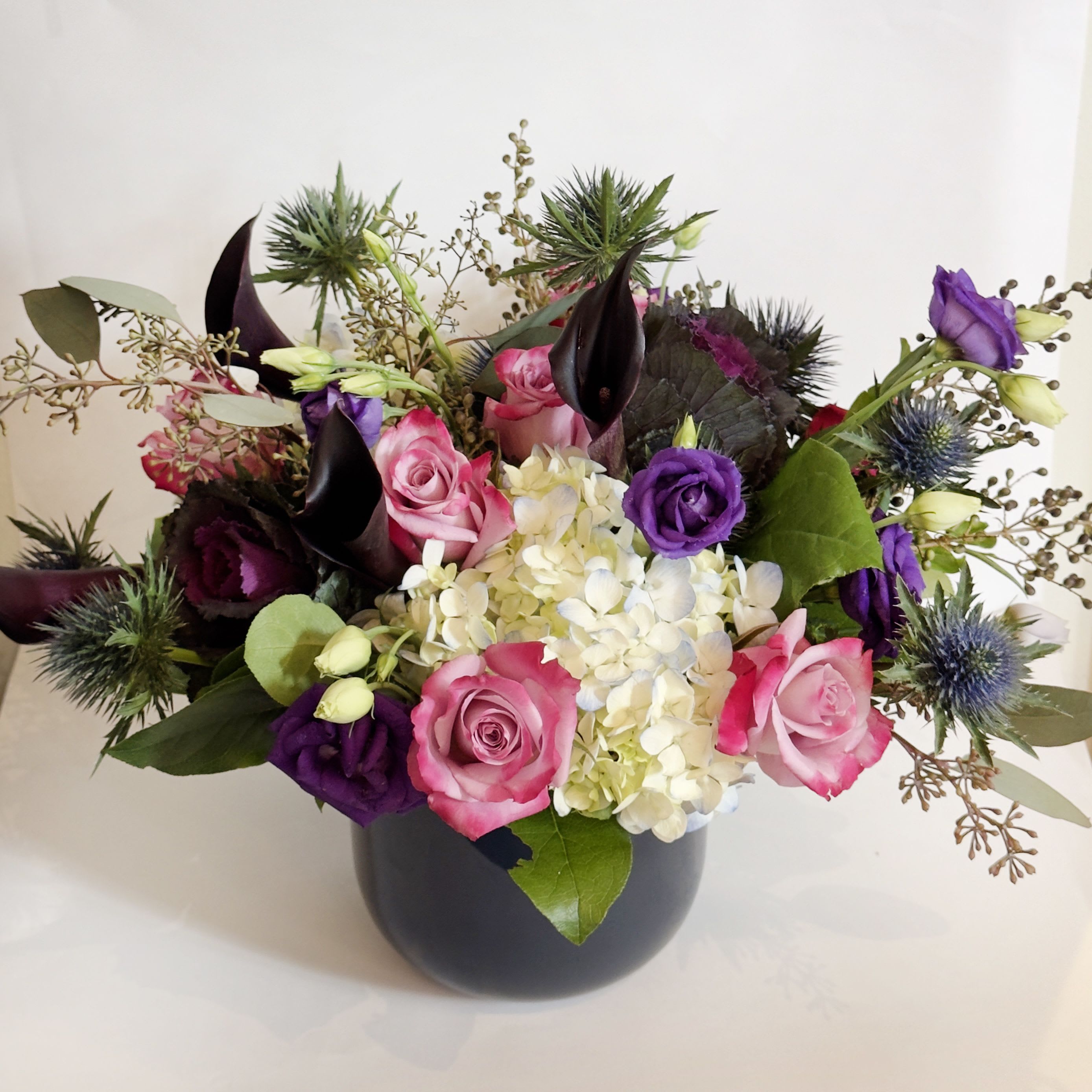 Rockfire Rockfire - This arrangement is presented in a dark blue glass vase. It feature dark purple kale, thistle, rockfire roses and blue hydrangea. It's a perfect late fall greetings.