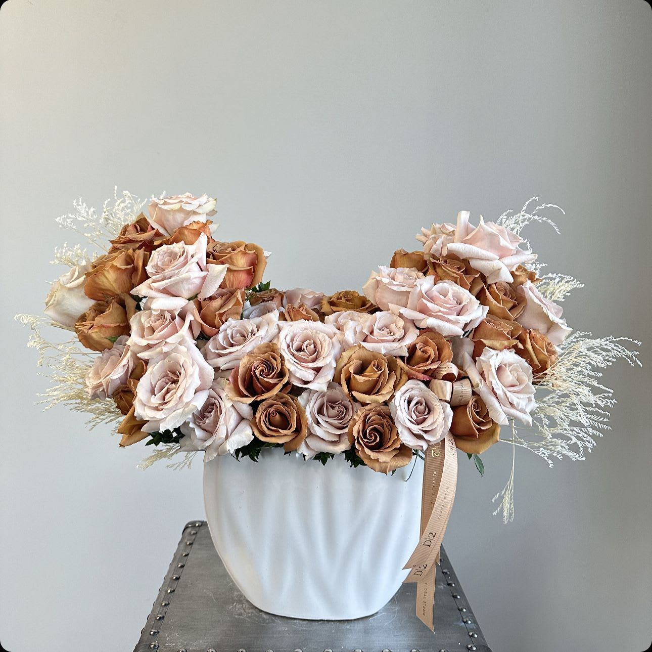 Brown Monica - This luxurious floral arrangement features a stunning blend of soft blush and golden-toned roses, artfully cascading in a balanced design. The muted tones are complemented by delicate, wispy white foliage, adding texture and an ethereal touch. Presented in a sleek, sculpted white vase, the arrangement exudes elegance and timeless sophistication, perfect for refined occasions or as a statement decor piece.