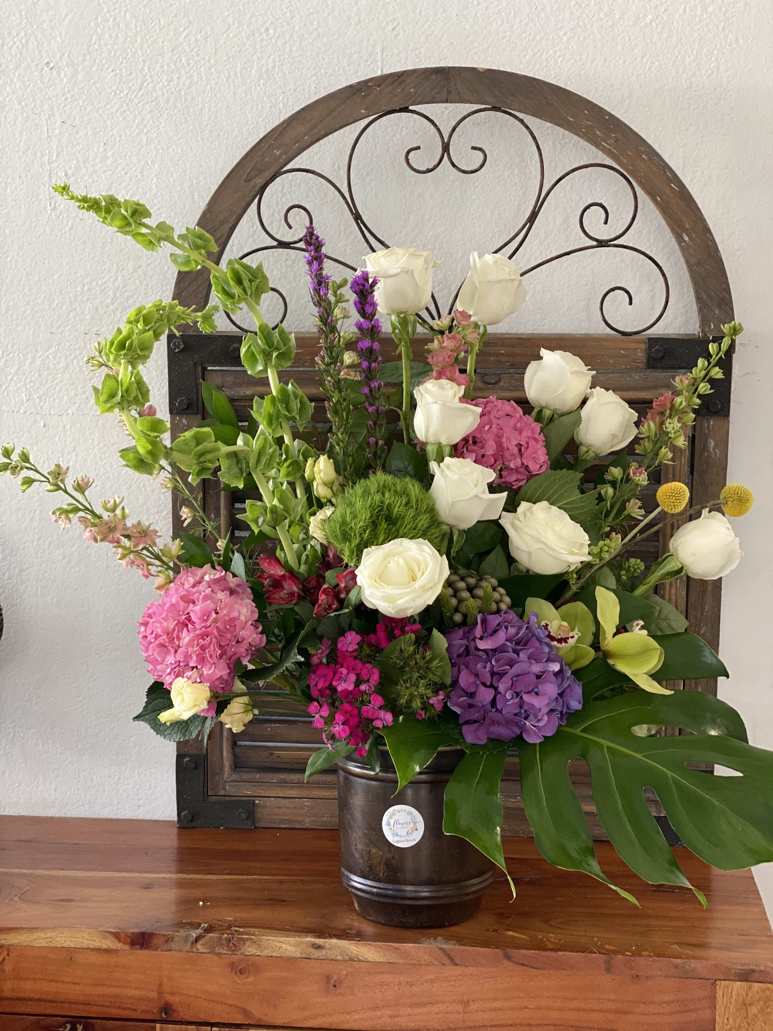 White goes with everything - Fun white roses with pink and purple flowers playing in tropical leaves and bells of Ireland arranged in a brown vase. 
