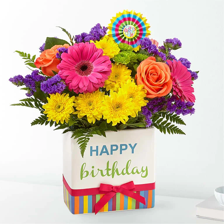 Birthday Brights Bouquet - The Birthday Brights Bouquet is a true celebration of color and life to surprise and delight your special recipient on their big day! Hot pink gerbera daisies and orange roses take center stage surrounded by purple statice, yellow cushion poms, green button poms, and lush greens to create party perfect birthday display. Presented in a modern rectangular ceramic vase with colorful striping at the bottom, &quot;Happy Birthday&quot; lettering at the top, and a bright pink bow at the center, this unforgettable fresh flower arrangement is then accented with a striped happy birthday pick to create a fun and festive gift.