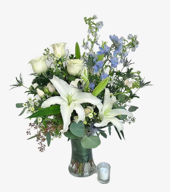 River of Dreams - Soft blue delphinium stands high above soft white blooms of roses and classic lilies, accented with light peach spray roses and textual blue thistle. Designed in a leaf-lined clear glass vase. Appropriate for delivery to services or home/office. 