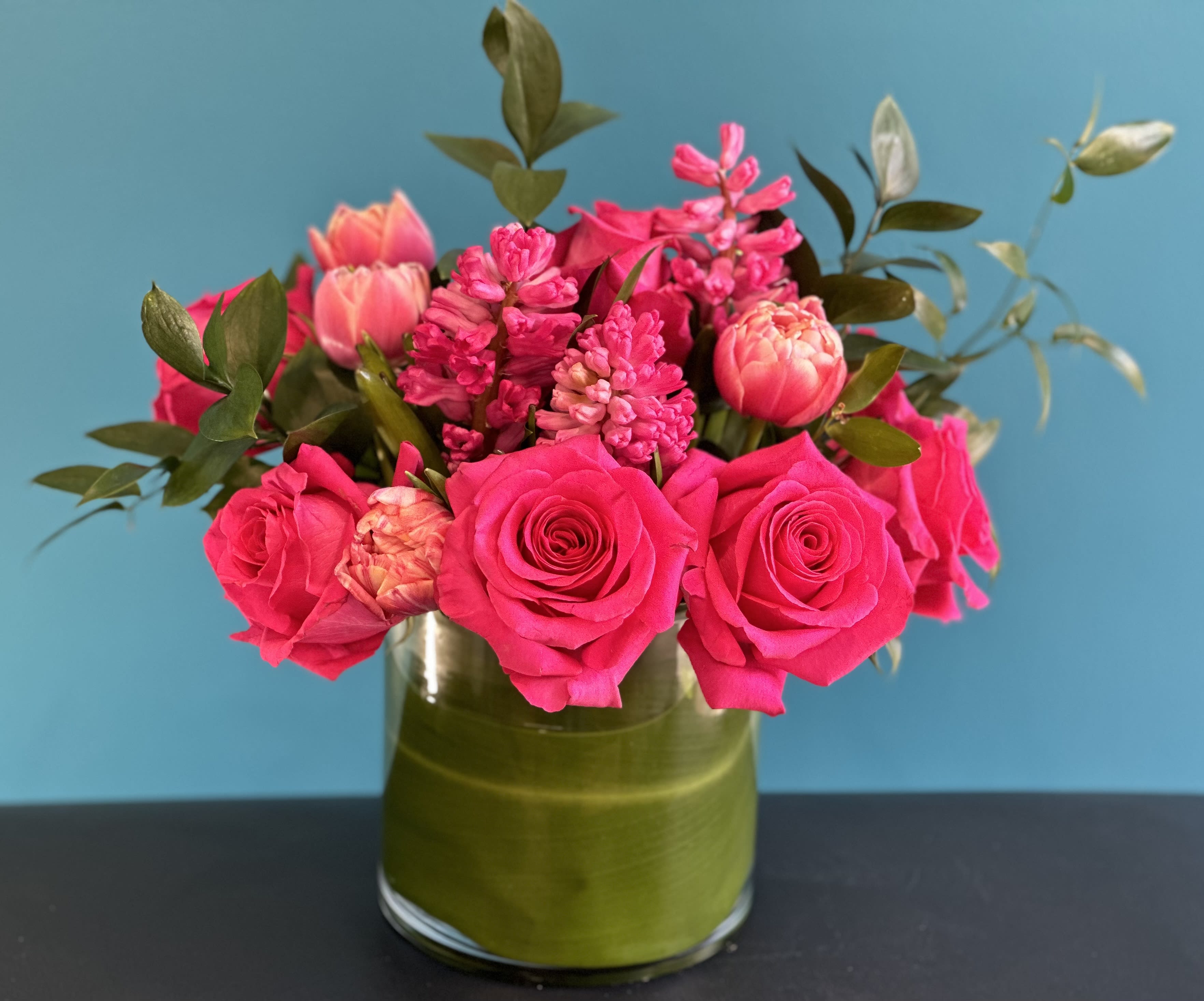 Hearts Desire Hearts Desire - Lush mix of hot pink roses, fragrant hyacinth, double pink tulip & mix of ruskus set in to a ti leaf lined 6" cylinder.
