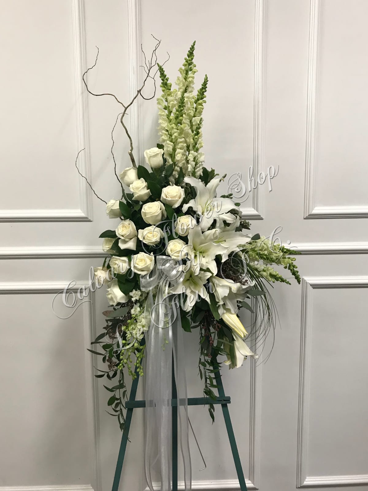 Adorned Condolences - A very unique and beautiful standing spray consisting of white roses, casa blanca lilies and white snapdragons; decorated with curly willow and cascading greens. 