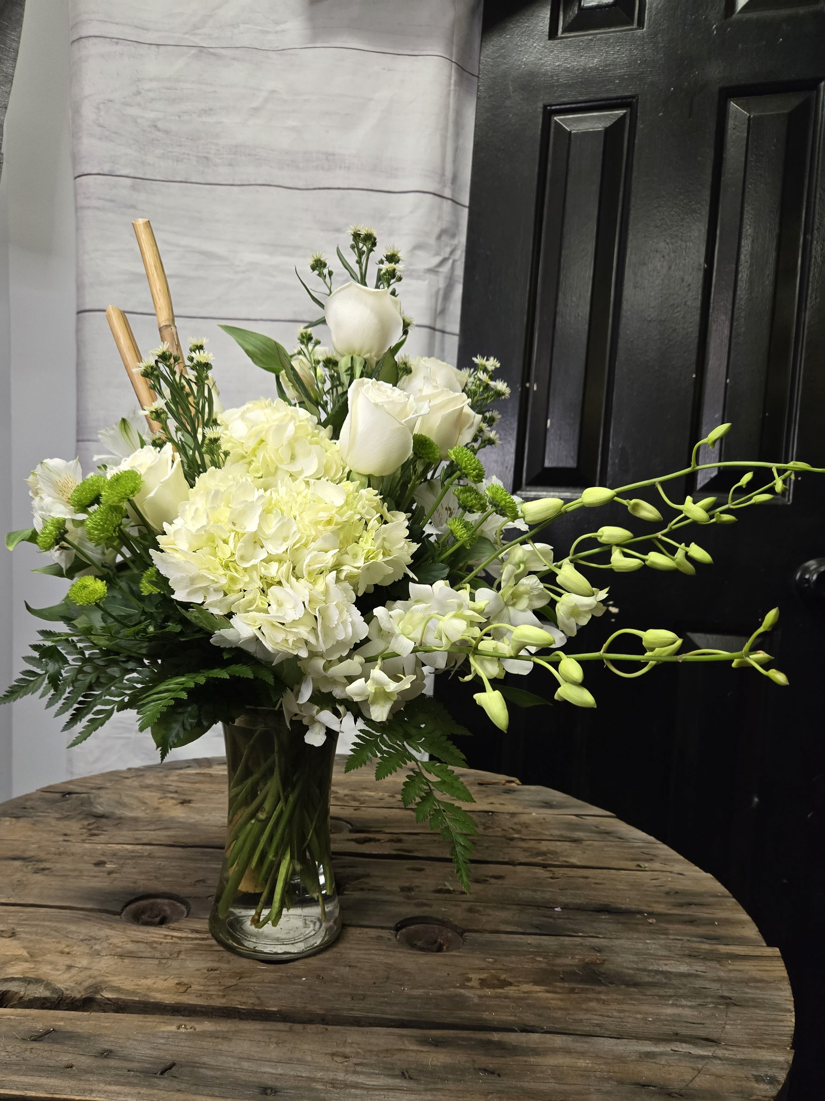 tranquility - This floral arrangement is an elegant and sophisticated design featuring a mix of white and green flowers, artfully arranged in a clear glass vase. The bouquet includes:  White Roses – Classic and timeless, these roses add a romantic and elegant touch. White Hydrangeas – Large, lush blooms that provide volume and softness to the arrangement. White Orchids – A cascading element with delicate buds and blossoms, adding grace and movement. Green Button Mums – Small, vibrant green flowers that add contrast and texture. Various Greenery – Ferns and other leafy foliage that frame the bouquet and enhance its natural beauty. Decorative Bamboo Sticks – A unique design element that adds height and an organic, structured look. The combination of soft white blooms and fresh green accents creates a fresh, luxurious, and tranquil aesthetic, making it suitable for various occasions such as weddings, sympathy arrangements, or elegant home deco