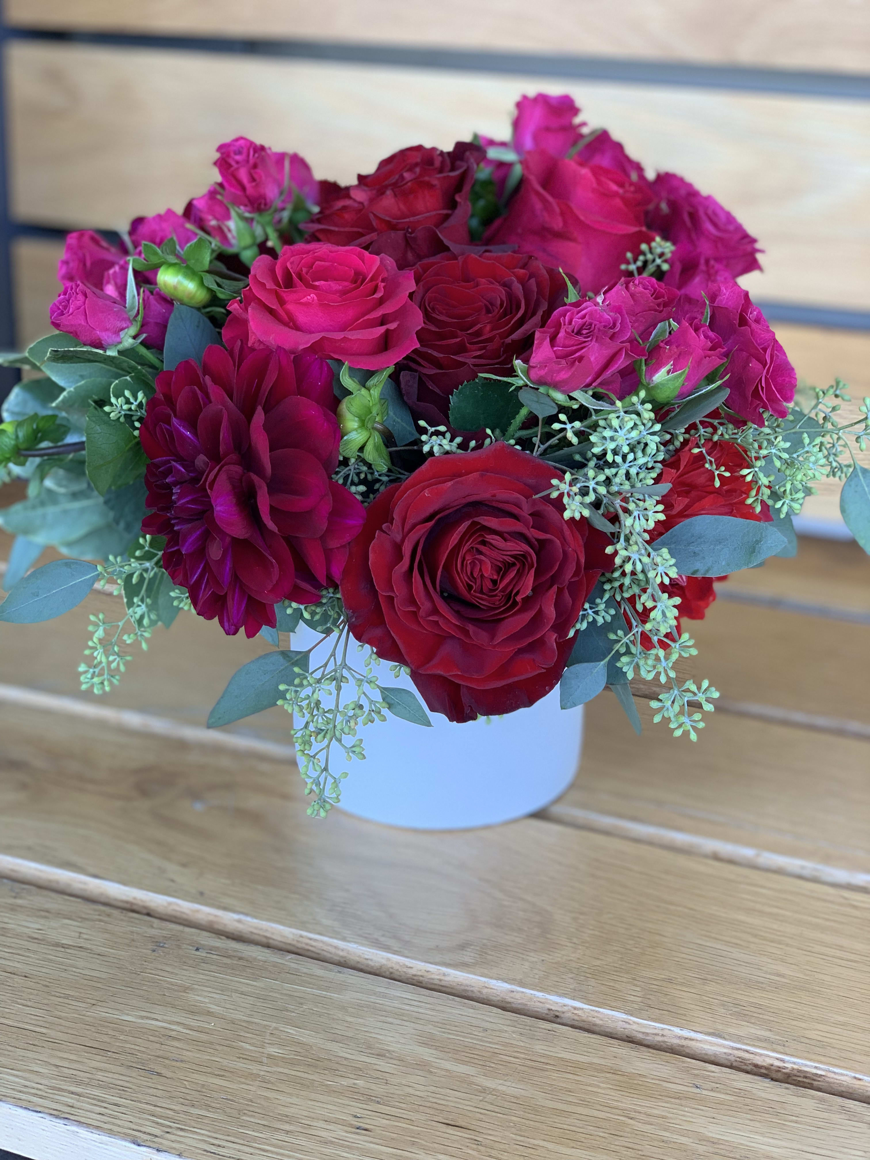 Red Velvet - Deep reds and hot pink roses, dahlias with a touch of seed eucalyptus arranged in a white ceramic vase.