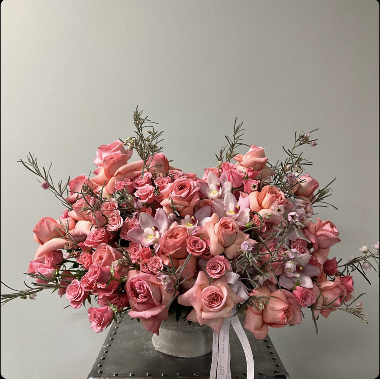Chloe Chloe - This is an elegant floral arrangement displayed in a simple, low vase. The arrangement features a variety of pink flowers, predominantly roses, interspersed with delicate orchids and small filler flowers. The bouquet is complemented by sprigs of greenery, creating a balanced and lush appearance. The soft, pastel tones of pink and peach add a romantic and sophisticated touch to the design. The vase itself is understated, allowing the vibrant blooms to remain the focal point.