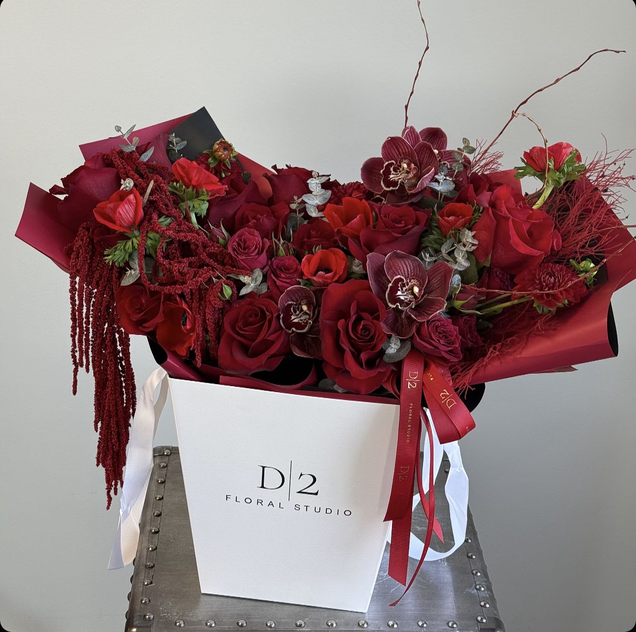 Madelyn Madelyn - This is a striking and dramatic bouquet presented in a sleek white box labeled "D2 Floral Studio." The arrangement showcases an opulent display of red roses, complemented by burgundy orchids, amaranthus, and other deep red and crimson blooms. Accents of eucalyptus and wispy branches add texture and depth, while the cascading amaranthus lends a luxurious and flowing effect. The bouquet is wrapped with rich red paper and ribbons, enhancing its bold and passionate aesthetic. This arrangement is perfect for a statement gift or a romantic occasion.