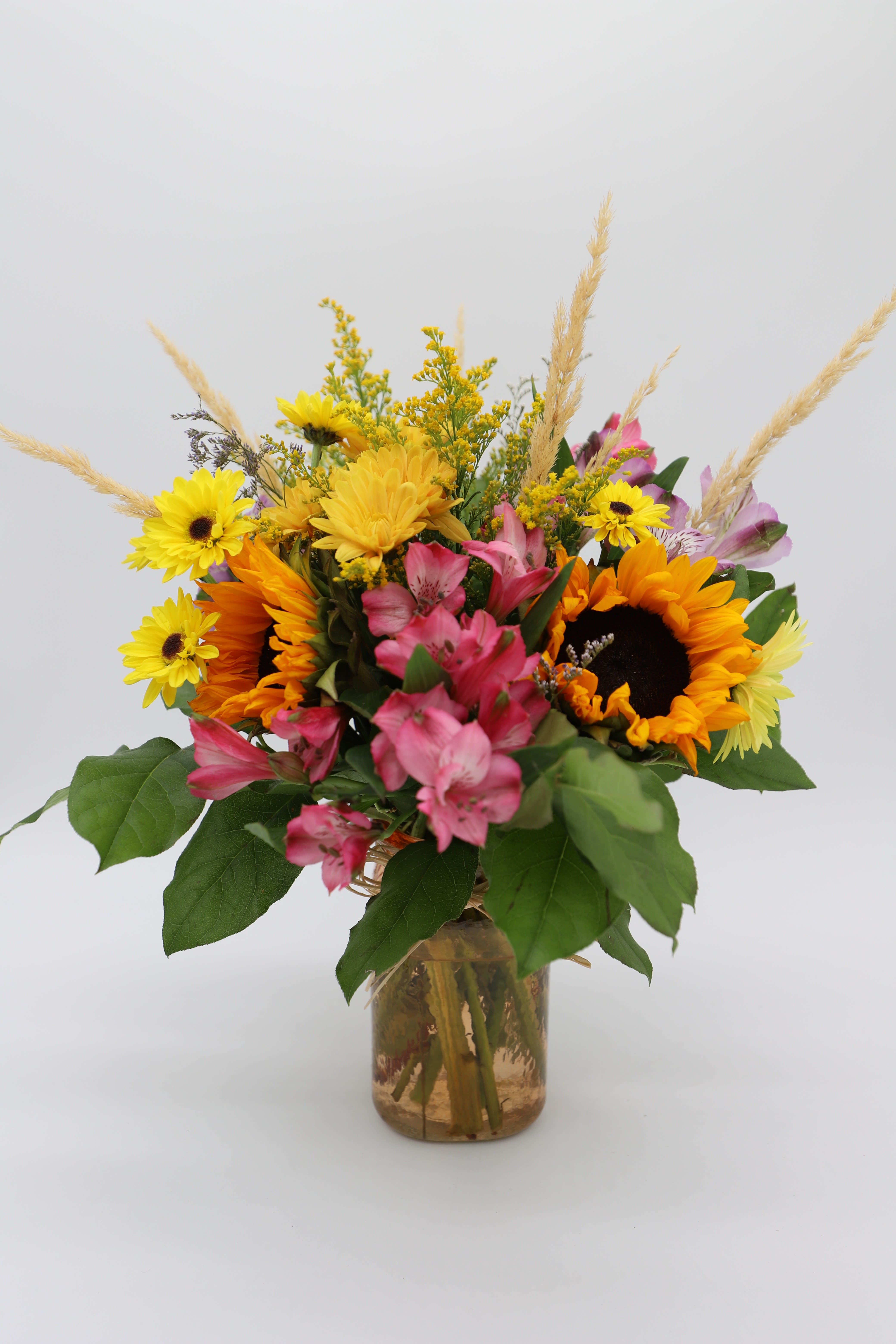 Glowing Harvest - An endearing floral arrangement to show your appreciation to any special person in your life! This arrangement has a beautiful bundle of sunflowers, alstromerias, mums, greenery and dried wheat stalk in a textured glass vase with a rafia bow.