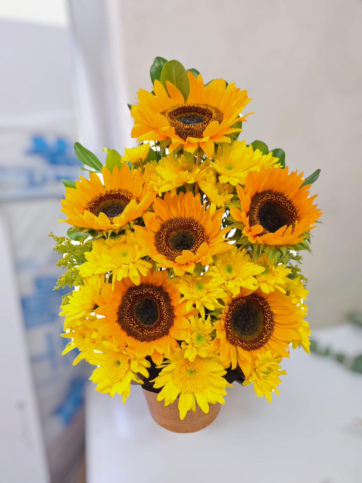 Ramo de girasol madre - Este ramo está compuesto principalmente por girasoles, con flores grandes de pétalos amarillos vibrantes y centros marrones oscuros. Los girasoles están dispuestos en una forma redondeada, creando un diseño compacto y lleno. Se aprecian algunas hojas verdes que complementan la composición. El fondo es borroso, lo que resalta aún más los colores brillantes del ramo. Es un arreglo alegre, perfecto para transmitir energía positiva y calidez.