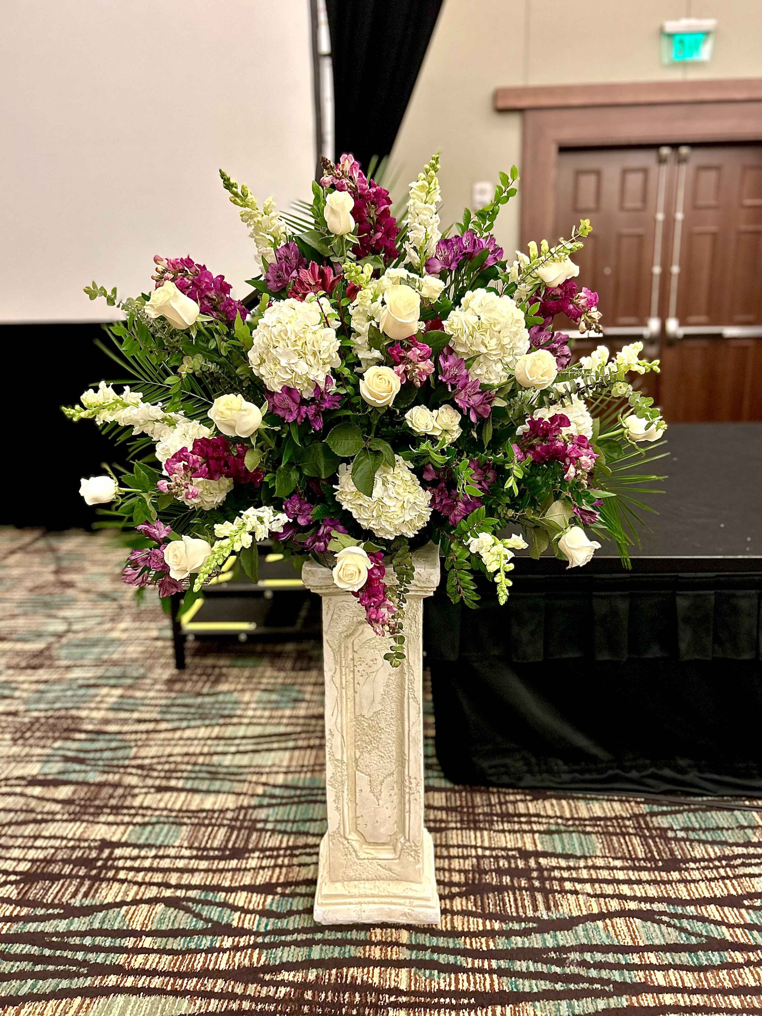 Elegance in Bloom Elegance in Bloom - A striking floral arrangement that captures grace and vibrancy, Elegance in Bloom combines the purity of white blossoms with the bold passion of fuchsia accents to add a touch of modern flair and exuberance. The flowers are carefully arranged to create a harmonious balance, with cascading greens and delicate filler blooms weaving through the composition. This stunning arrangement sits atop a pedestal at the head of the podium, exuding sophistication and beauty for the ceremony, symbolizing new beginnings and vibrant futures.