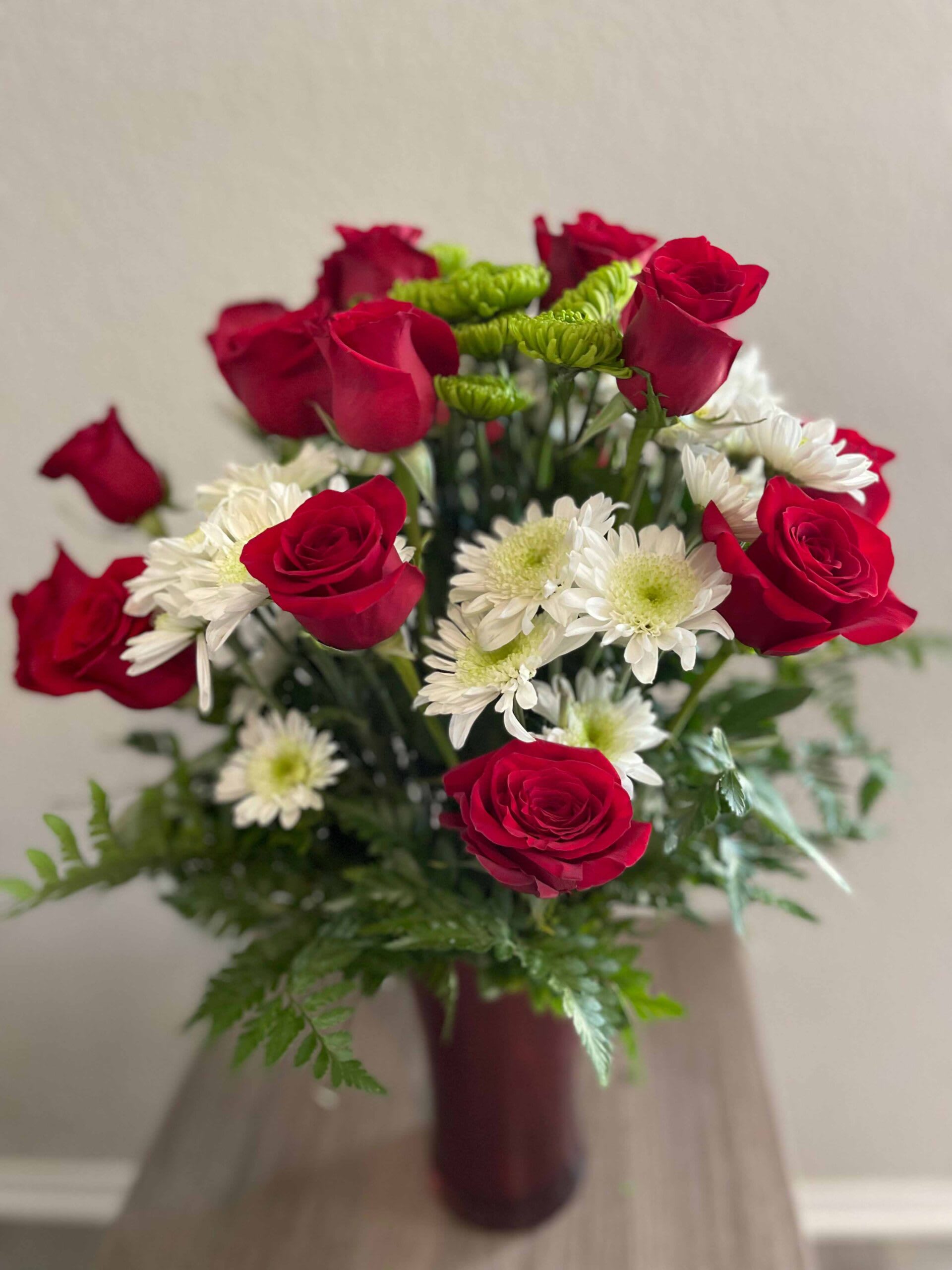 Blossom Stories - Beautiful  floral arrangement of  18 long stem red roses, white and green mums and leather leaves all around on a tall red vase.