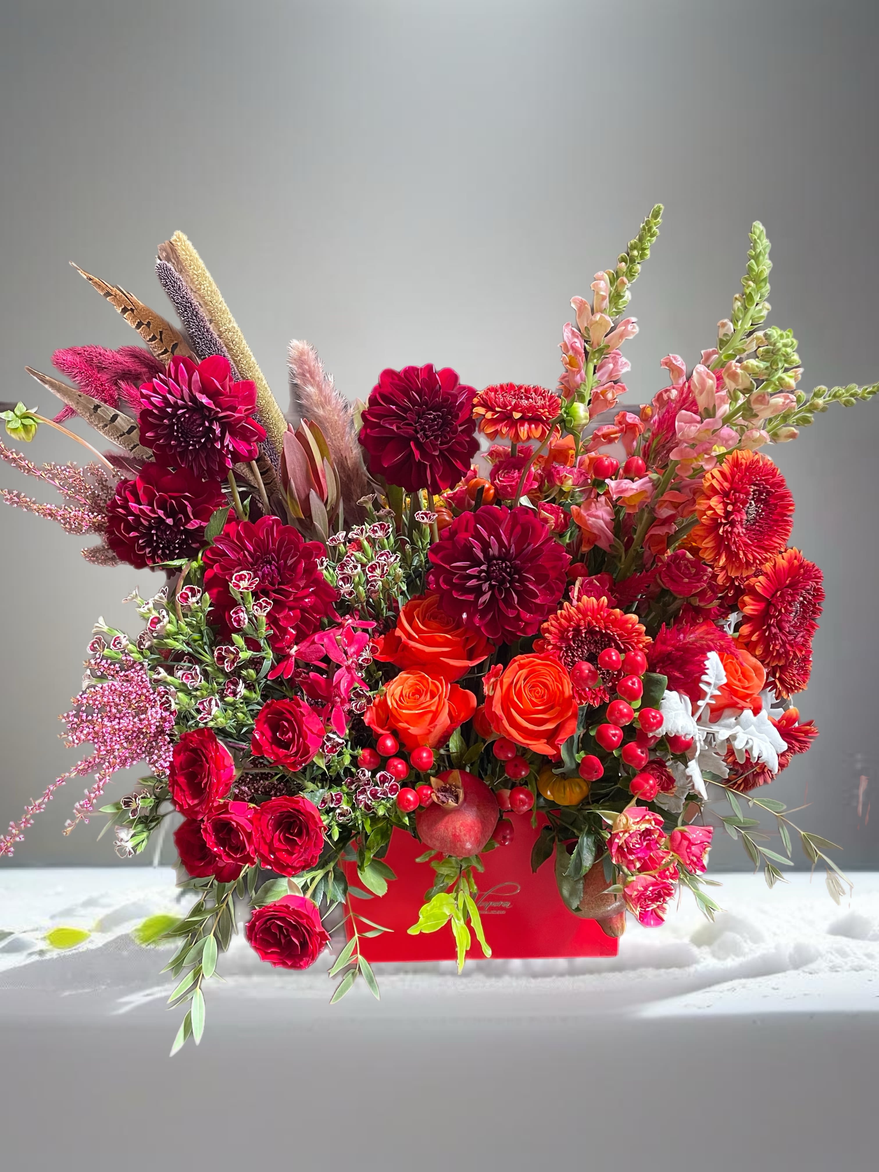 Rococo Boho in Red  - Introducing our Rococo Red Flower Arrangement, elegantly presented in a vibrant red gift bag. This stunning display features various shades of red roses, dahlias, chrysanthemums, and gerbera flowers, enhanced by soft feathers and boho elements. Accented with pomegranate and mini pumpkins, it's a perfect centerpiece for Thanksgiving. For added festivity, we can include seasonal fruits like grapes, apples, or strawberries. This arrangement is a beautiful blend of drama and warmth, ideal for any celebration!