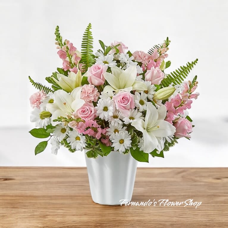 Divine Confort - This charming floral arrangement exudes softness and grace, making it a perfect expression of love and remembrance. Featuring a delicate mix of pastel pink roses, carnations, and snapdragons paired with elegant white lilies and daisies, this bouquet symbolizes purity, affection, and peace. Accented with lush greenery and arranged in a classic white vase, it radiates natural beauty and serenity. Ideal for honoring cherished memories or sending heartfelt sentiments, this design offers a comforting and uplifting gesture.
