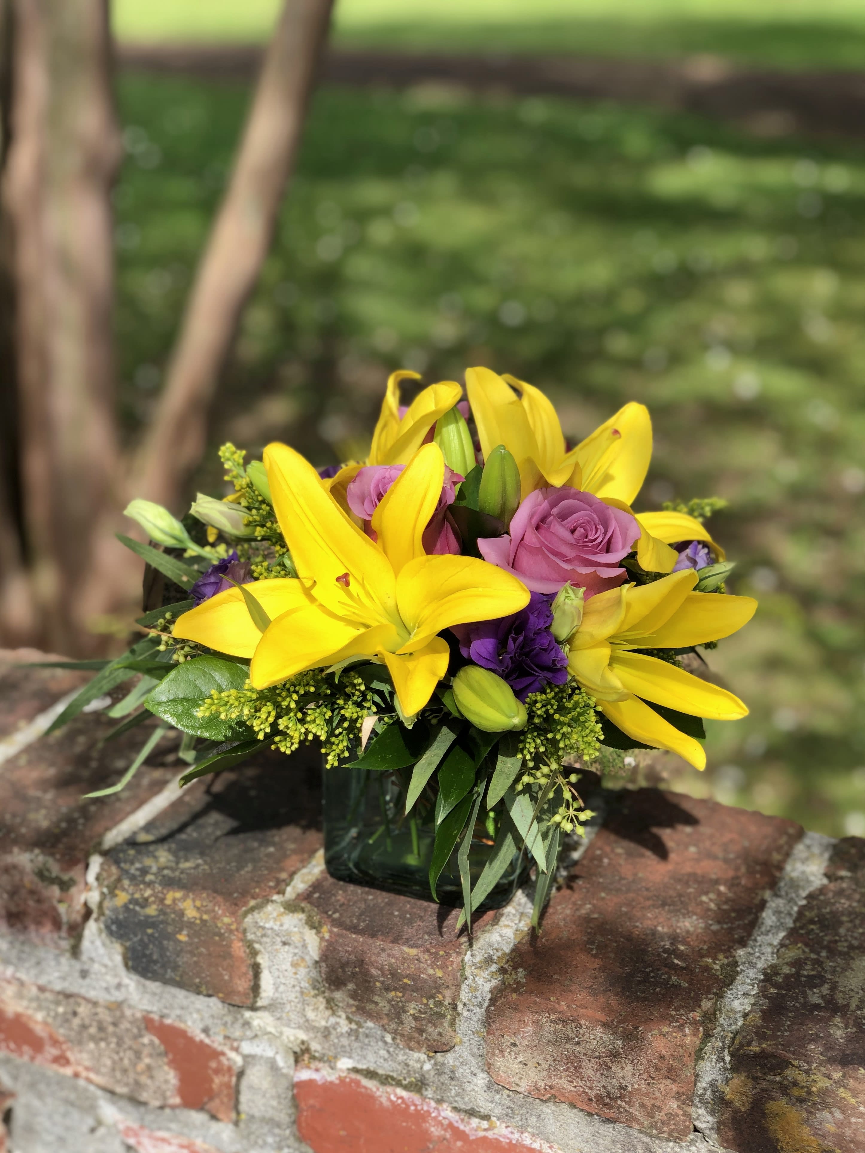 High Impact - Make a statement with this elegant arrangement! Created in a simple square vase with yellow Asiatic lilies accented by shades of lavender and purple roses carnations and fragrant aspidistra this arrangement is bound to make an impact. When you need to impress sending this high energy flower arrangement leaves a lasting impression.