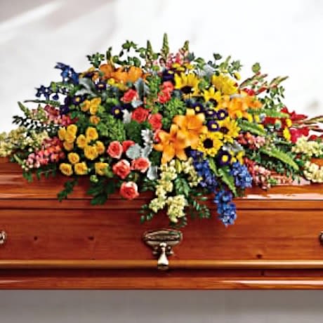 Eternal Rest - The image shows a beautifully decorated casket adorned with a vibrant floral arrangement. The flowers are a mix of colorful blooms, including yellows, oranges, blues, reds, and greens, creating a striking and lively display. This arrangement features a variety of flowers such as roses, lilies, daisies, and other seasonal blossoms, interspersed with lush greenery. It appears to be a classic tribute, often used for memorial or funeral services, symbolizing respect, love, and remembrance. The casket is a polished wood design, adding a touch of elegance to the setting.