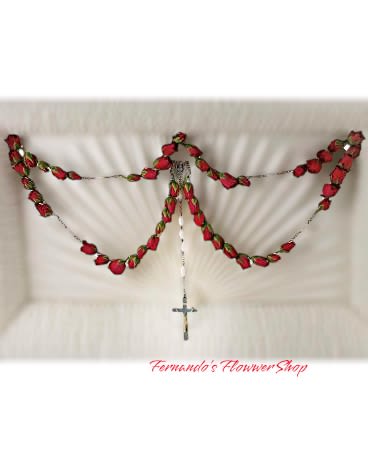 My Last Mystery My Last Mystery - The image displays a delicate rosary design crafted from red roses, arranged in the shape of a traditional rosary. The arrangement features interconnected rosebuds forming the beads of the rosary, draped gracefully against a white background, possibly the interior of a casket. At the center hangs a silver or metallic cross, completing the rosary design. This floral piece is a deeply religious and symbolic tribute, often used in Catholic funerals to honor the faith and memory of the deceased. The arrangement is elegant and spiritual, conveying reverence and devotion.