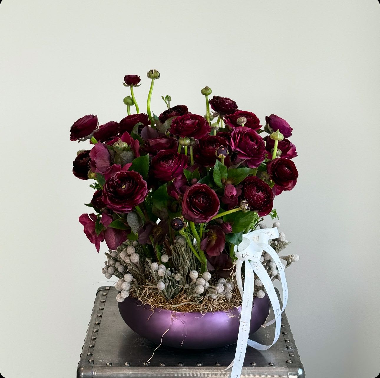 Layla Layla - This is an elegant floral arrangement featuring deep burgundy ranunculus blooms, complemented by lush green foliage. The flowers are artistically arranged in a sleek, metallic purple bowl, accented with soft white berries and moss at the base. A delicate ribbon adds a touch of sophistication, making this display perfect for formal occasions or as a statement piece in elegant settings.