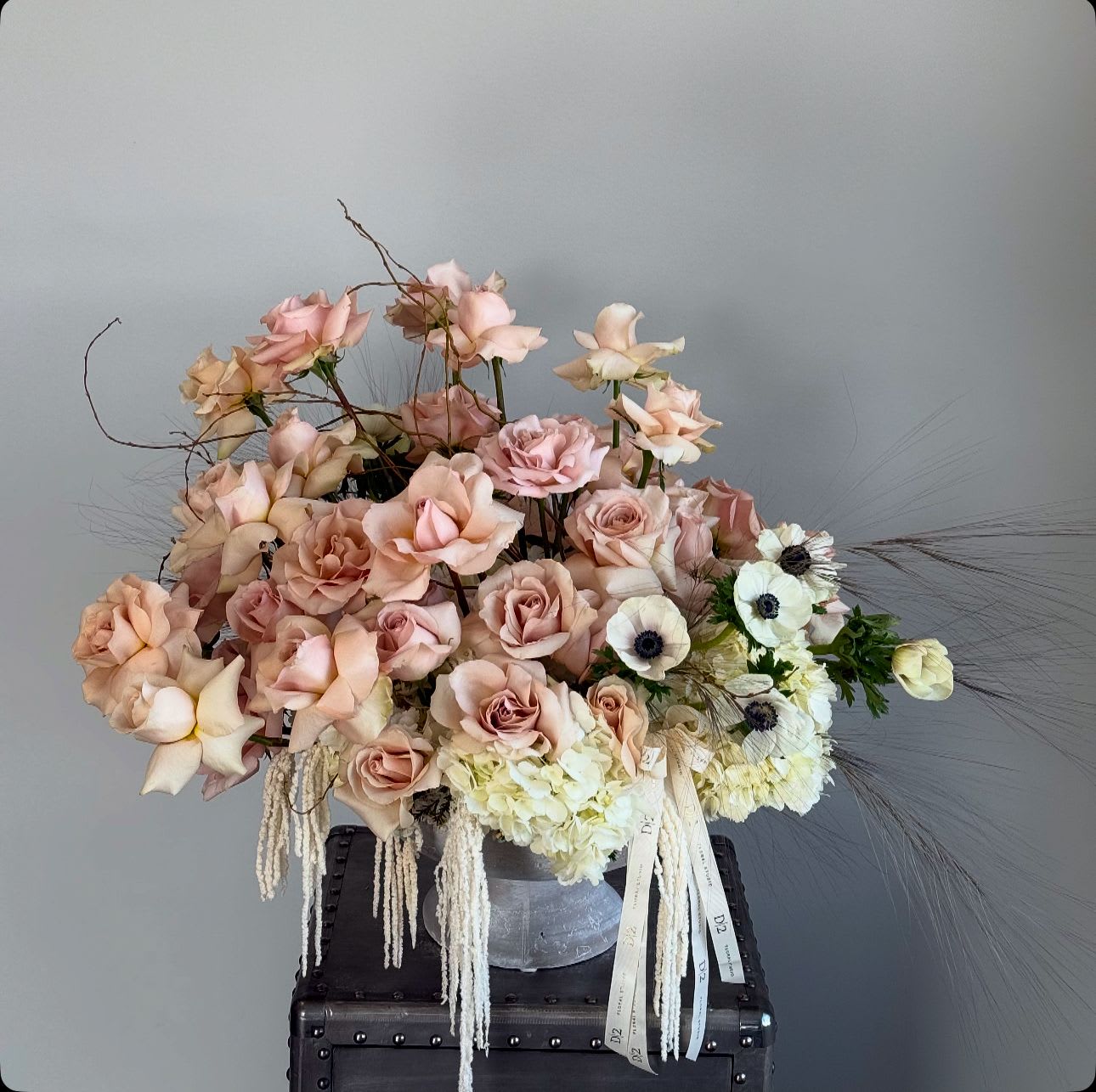 Zoe - This is a delicate floral arrangement featuring soft blush roses, creamy hydrangeas, and white anemones with dark centers. The arrangement is styled in a light, airy manner, enhanced with trailing amaranthus and subtle branches for a natural, organic feel. It is placed in a translucent vase with a ribbon accent, creating an elegant and romantic design perfect for weddings, formal events, or as a luxurious centerpiece.