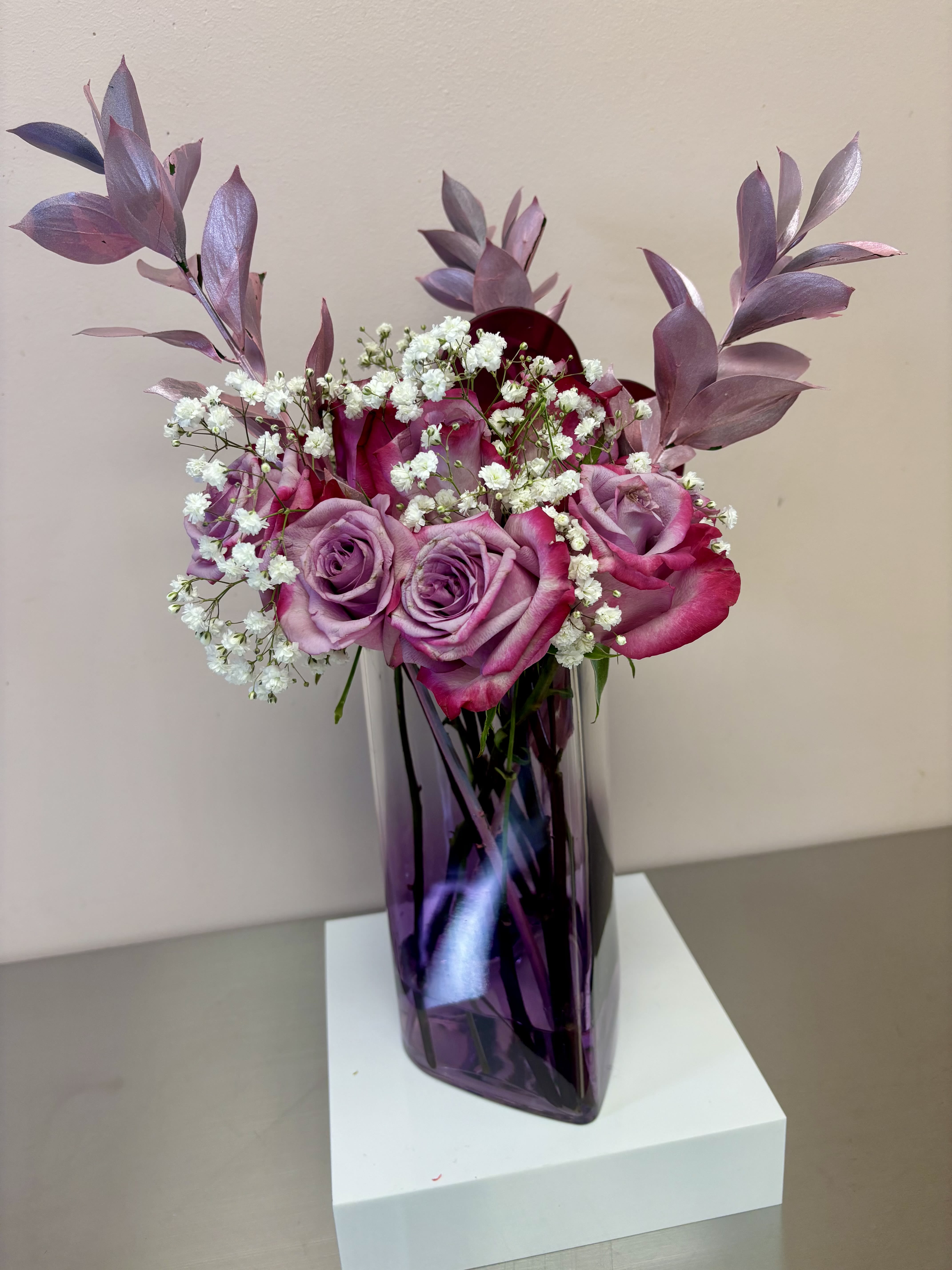 Rose Quartz - This charming floral arrangement features a modern aesthetic with its sleek, violet-hued 60-ounce heart-shaped canister and a beautifully curated bouquet of deep purple roses, accented with delicate baby’s breath for a touch of softness. The arrangement is elevated with striking tinted ruscus, adding a contemporary and elegant flair.  The vibrant colors and reflective accents create a perfect balance of boldness and sophistication, making it an ideal centerpiece for a modern space or a thoughtful gift to brighten someone’s day. 
