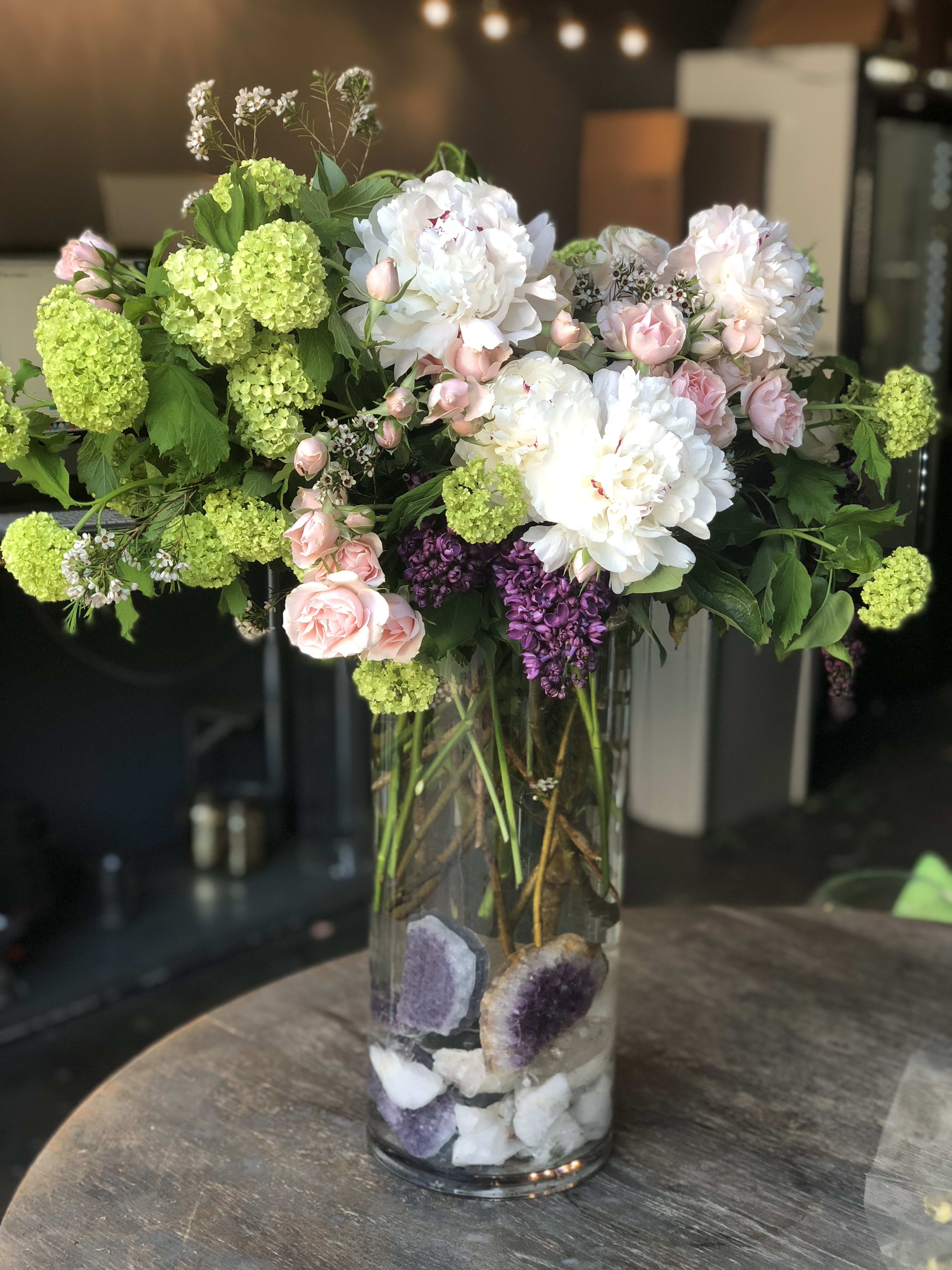 Bella Sensazione - Good feels with this grand design featuring healing stones like amethyst, quartz and more. This design includes peonies, lilac ( when in season) garden roses, spray roses, freesia and more in a 24 inch glass cylinder. 