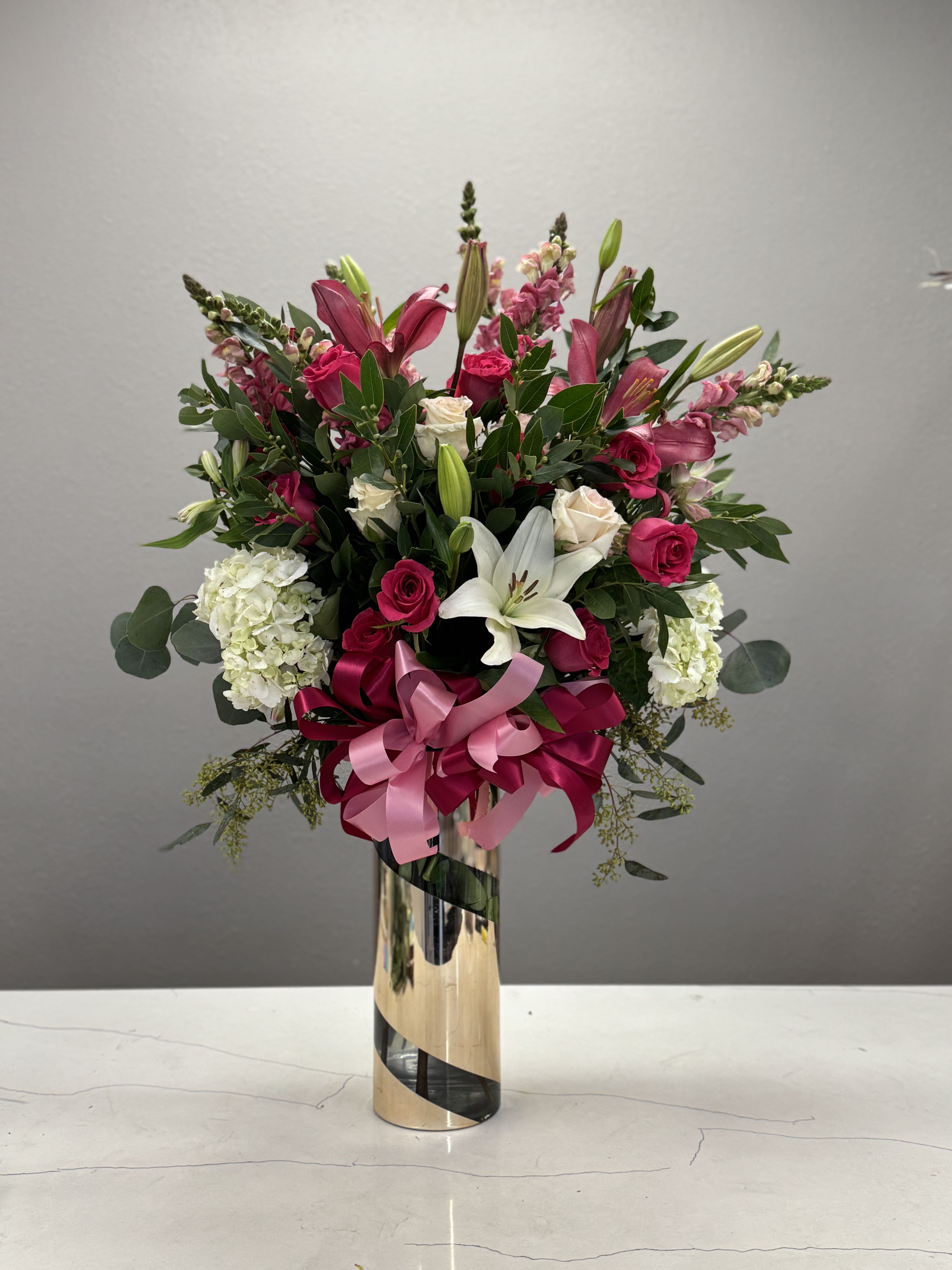 Elegant Blush - Add a touch of luxurious elegance to any space with this stunning floral arrangement. Featuring a vibrant mix of hot pink roses, delicate white hydrangeas, and bold hot pink and white lilies, this design radiates beauty and charm. All blooms are arranged artfully in a sophisticated gold tall vase, complemented with a soft pink ribbon for an added touch of grace. Perfect for any celebration, romantic setting, or to brighten your home with an unforgettable burst of color. 