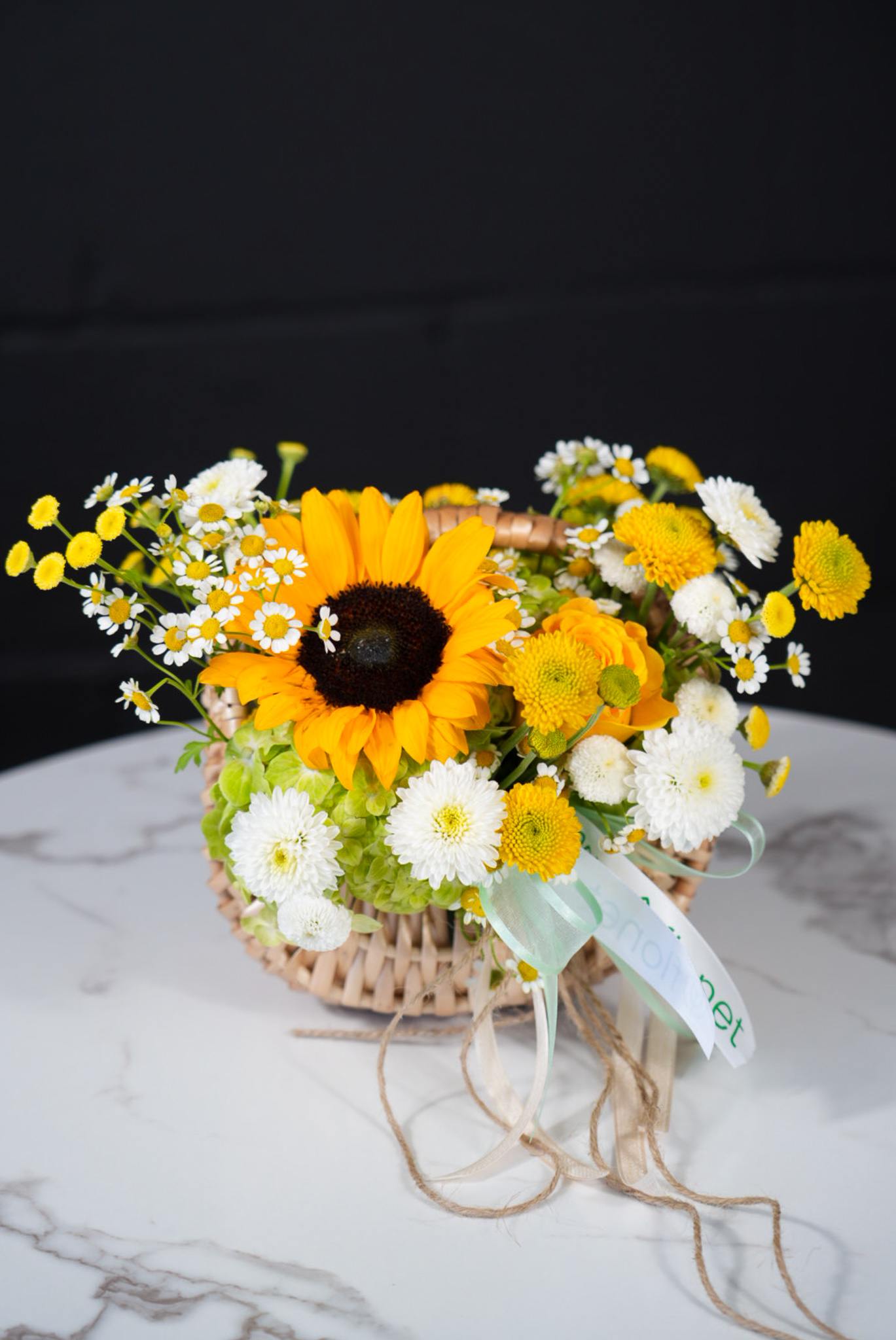 Flonet Petit Sunshine Basket - Brighten up any space with this radiant Sunshine Bloom Basket, a charming arrangement featuring a vibrant sunflower as the focal point, surrounded by delicate white and yellow chrysanthemums, playful chamomile flowers, and lush green hydrangeas. Designed in a rustic woven basket with soft ribbon details, this arrangement exudes warmth and happiness, making it a perfect gift for birthdays, celebrations, or simply to bring joy to a loved one’s day.